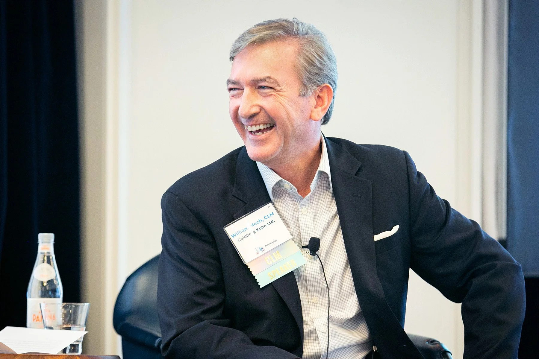A middle-aged man with gray hair, wearing a black suit and white checkered shirt, smiling during a meeting or conference. He has a name tag on his suit that reads 'William Mech, CLM, Goldman & Kohn Ltd.' with some additional badges or ribbons attache