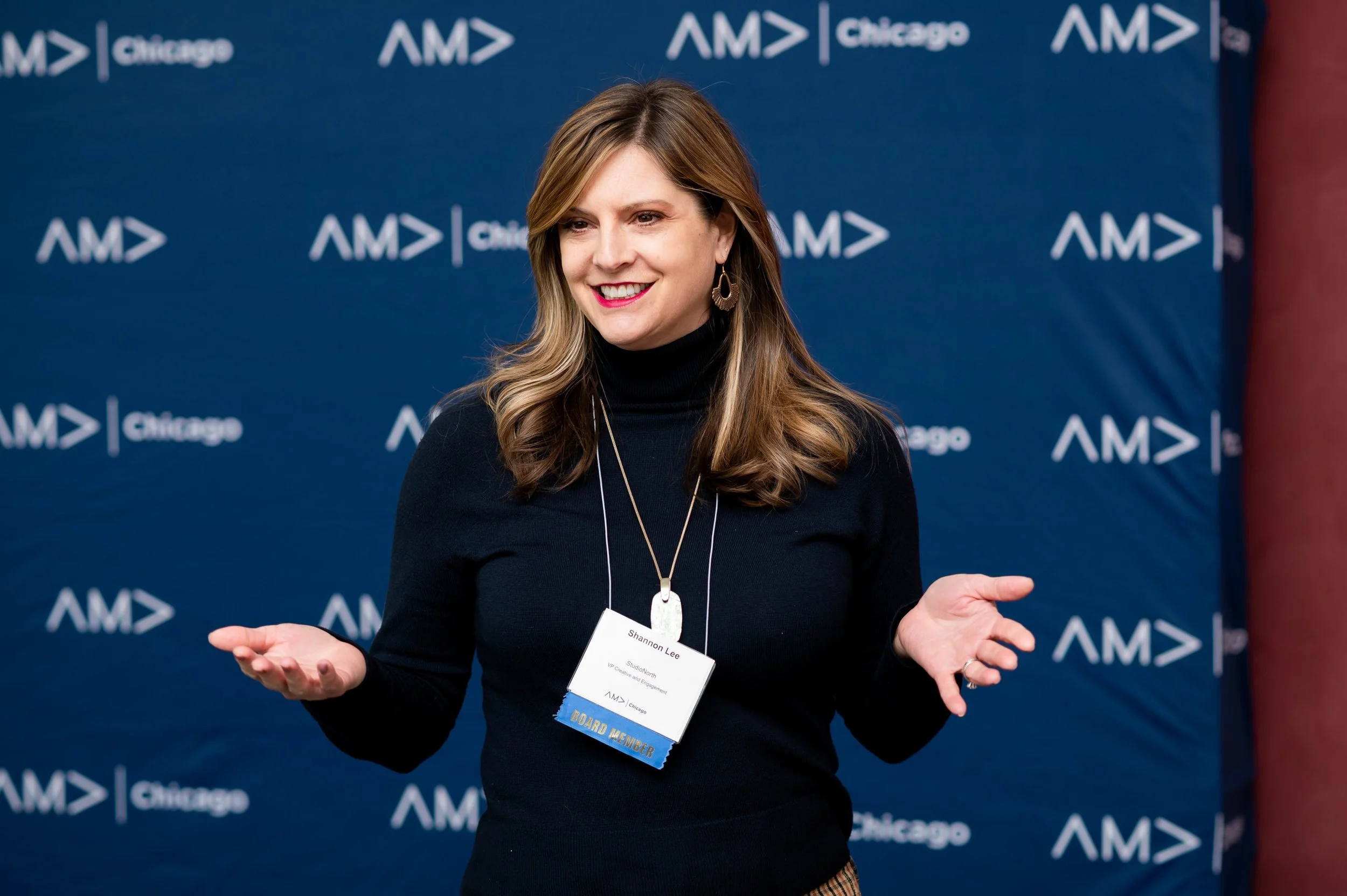 A woman with long hair smiling and gesturing with her hands at a conference, wearing a black turtleneck, with a name tag that reads "Shannon Lee" and a blue lanyard, standing in front of a blue backdrop with white logos that say "AM  Chicago."