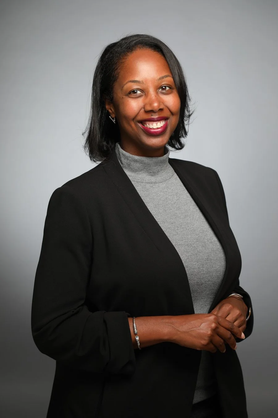 A professional woman with dark hair, smiling, wearing a black blazer over a gray turtleneck, and earrings, standing in front of a plain gray background.