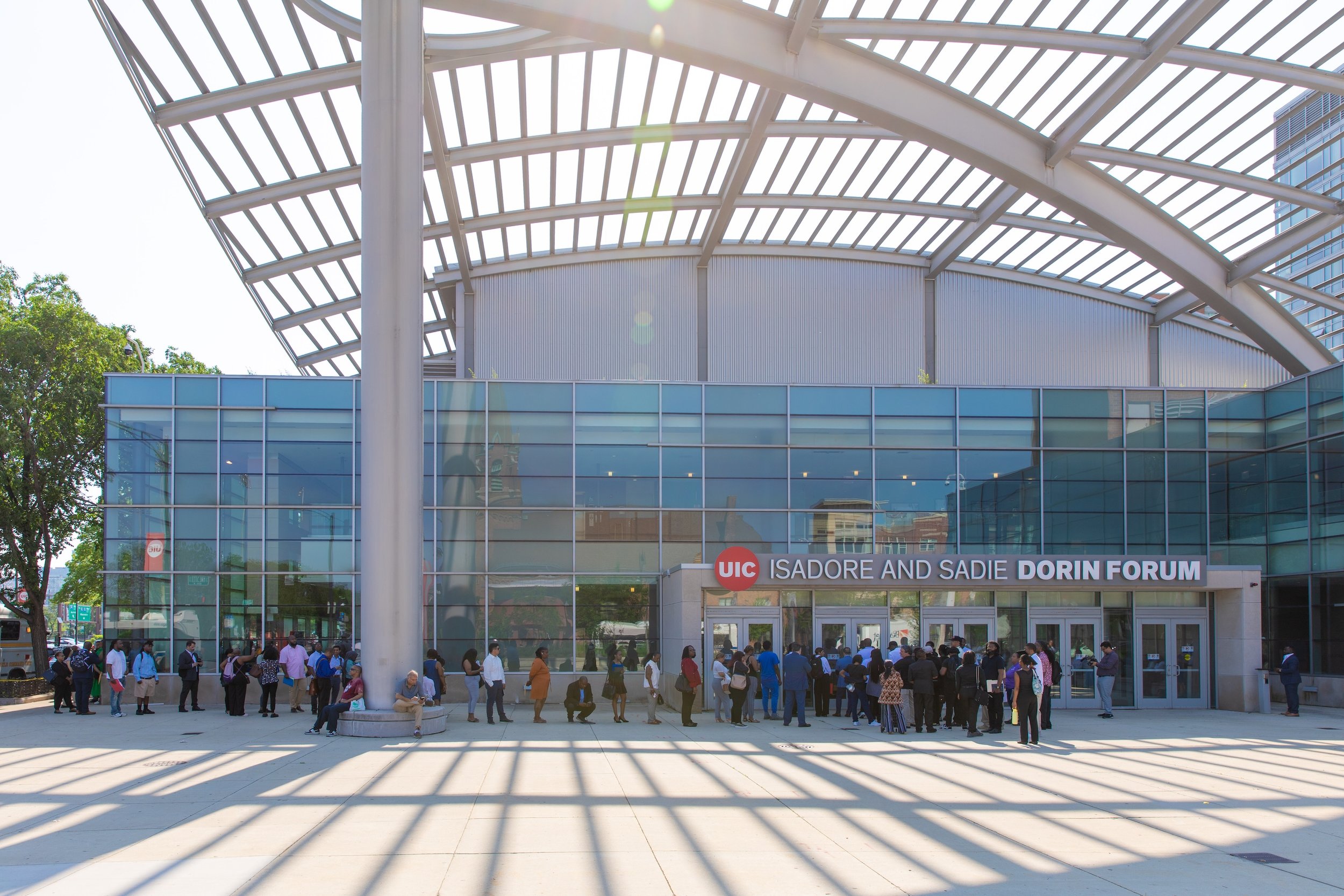 Crowd of people waiting outside the Isadore and Sadie Dorin Forum at the University of Illinois at Chicago, with a modern glass and metal building in the background and sunlight casting shadows on the tiled plaza.