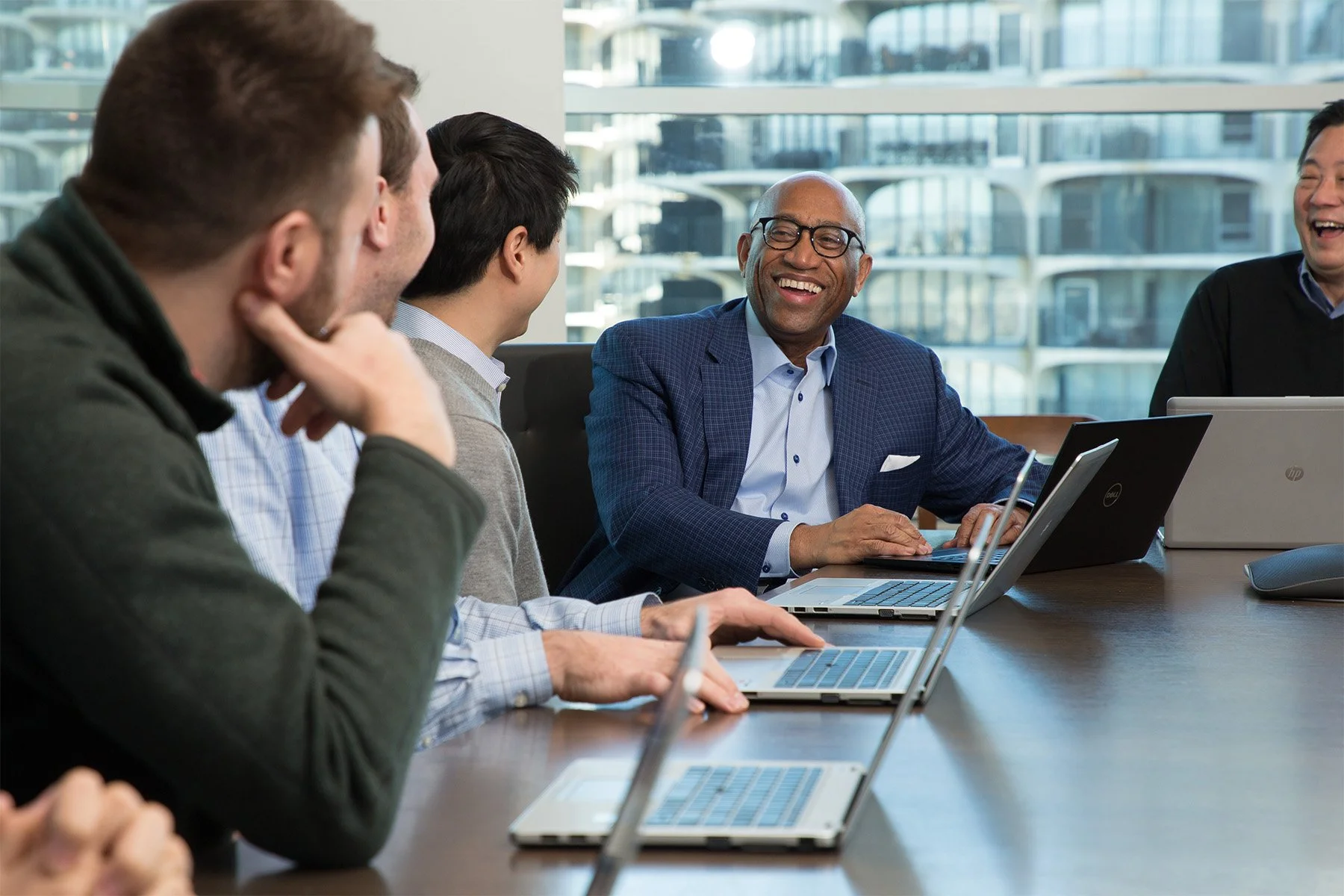 A diverse group of business professionals sitting at a conference table during a meeting, using laptops and engaging in conversation, with a cityscape visible through large windows in the background.