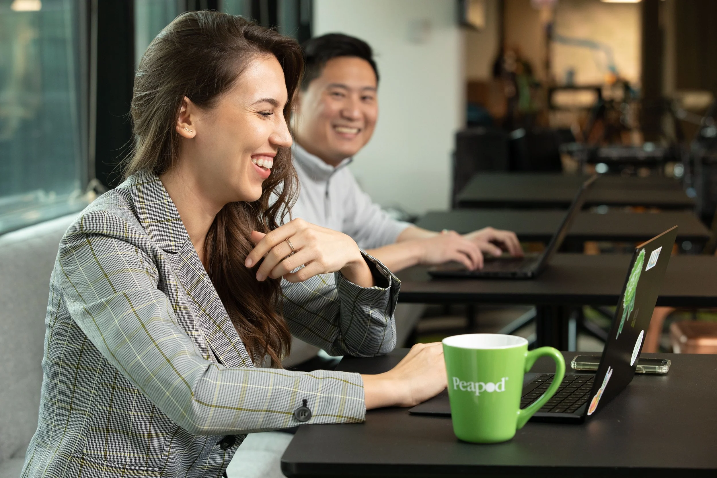 Two people working on laptops at a table, smiling and engaging, with a green mug labeled 'Peapod' in the foreground.