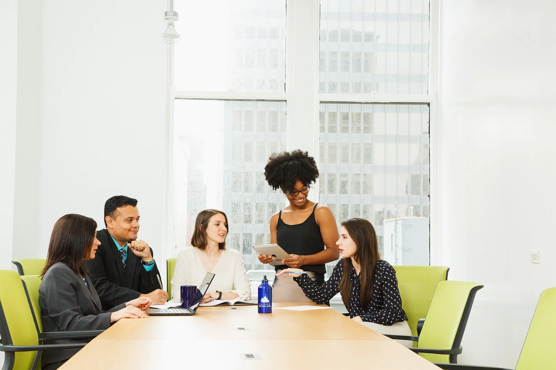 A diverse group of five people in a business meeting in a bright conference room with large windows, some seated and one standing and speaking while holding a tablet.
