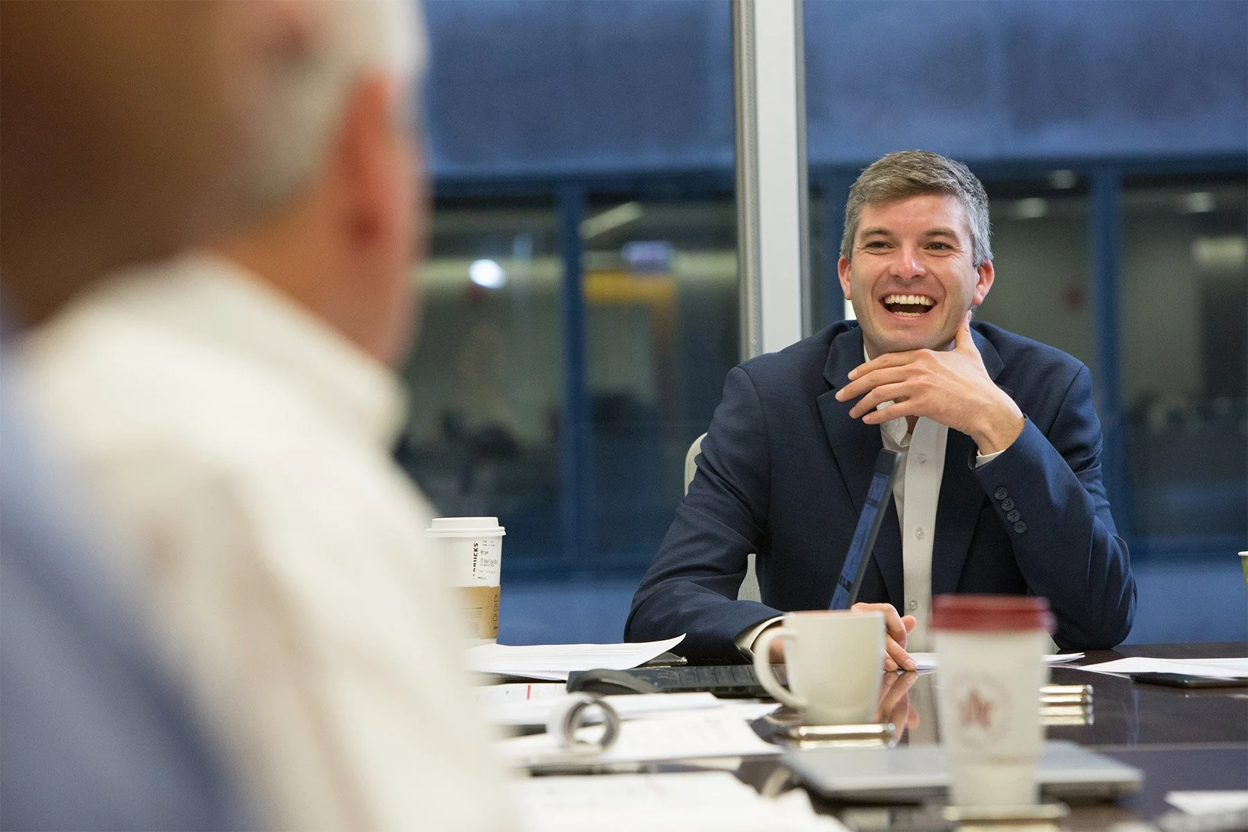 Two men in a business meeting sit at a conference table. One man in focus is smiling, wearing a dark suit and white shirt. The other man, out of focus, is seen from behind. The table has papers, coffee cups, and a laptop.