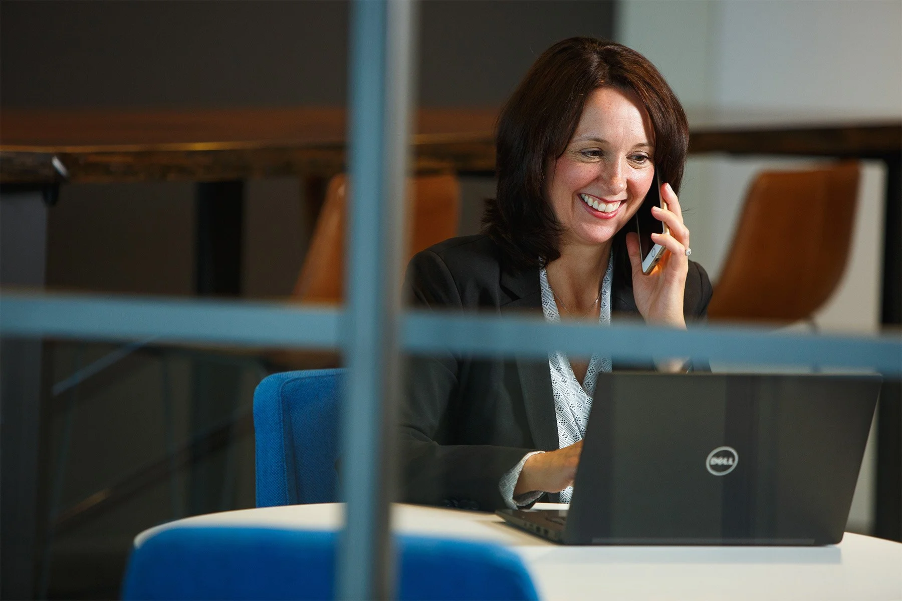 A woman with shoulder-length dark hair smiling and talking on her cellphone while working on her laptop in an office.