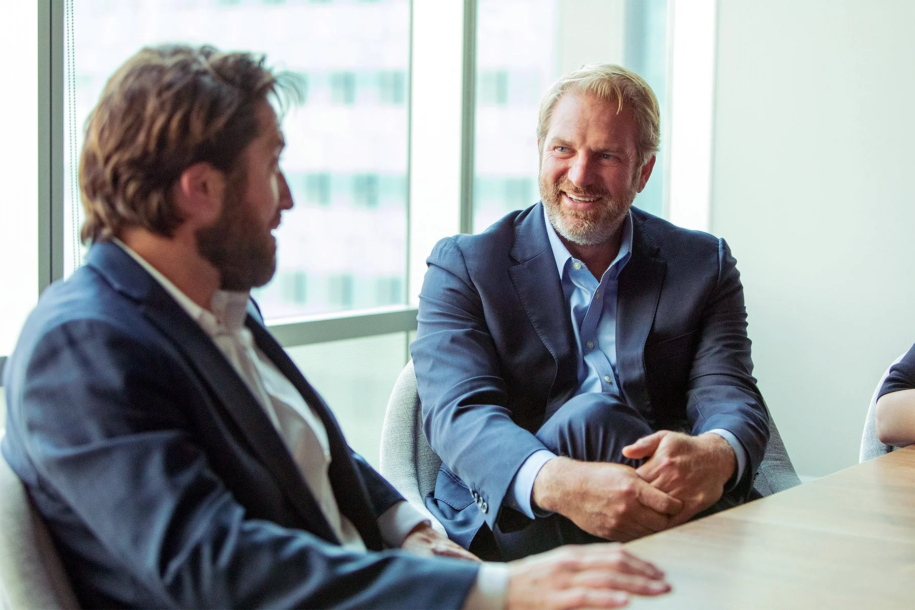 Two men in business suits sitting at a conference table, engaging in conversation, with a large window in the background.