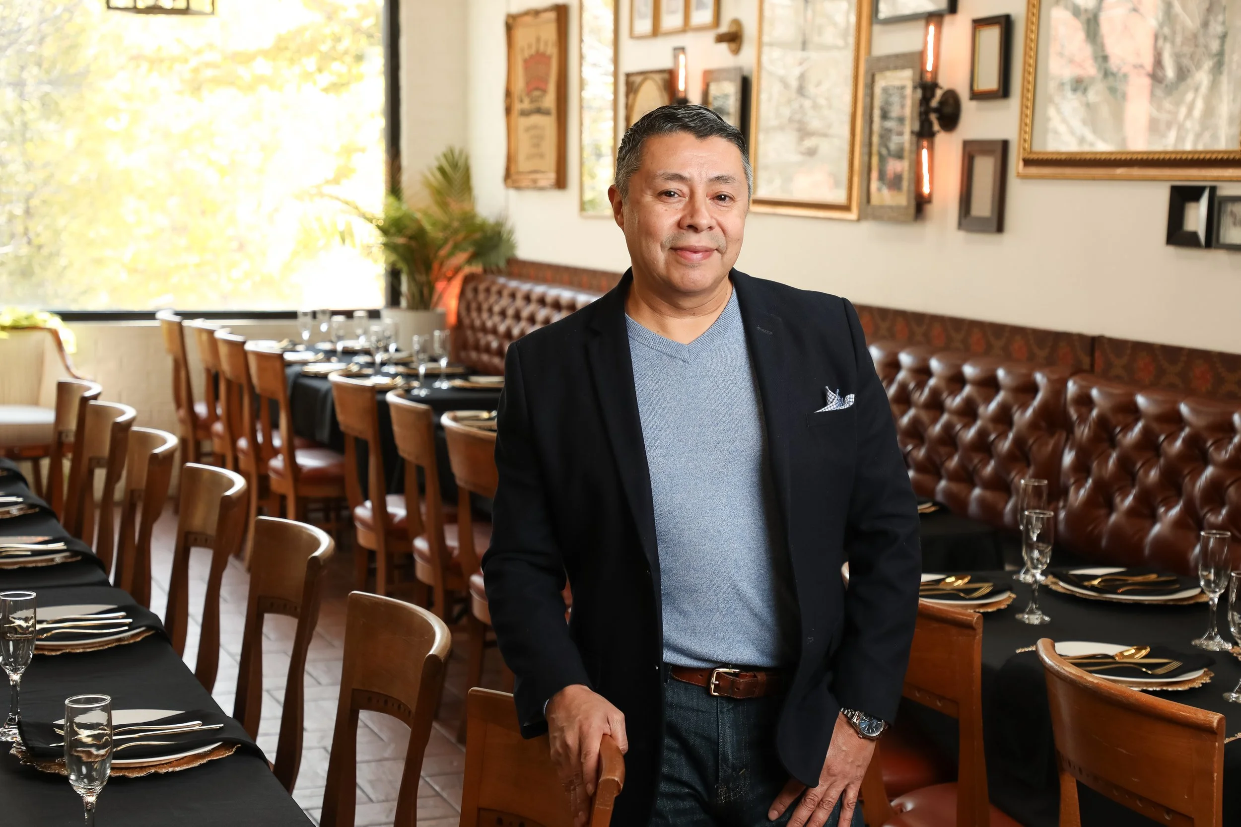 A man in a dark blazer and gray sweater standing in a restaurant dining area, with tables set with black tablecloths, glassware, and gold cutlery, and decorated with framed artwork and plants on the wall.