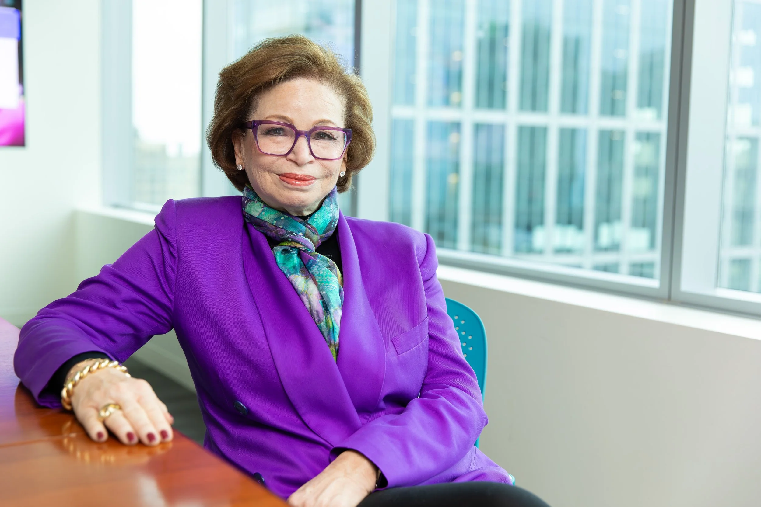 A woman with short brown hair, wearing purple glasses, a purple blazer, and a colorful scarf, sitting at a table in a modern office with large windows and a cityscape view.
