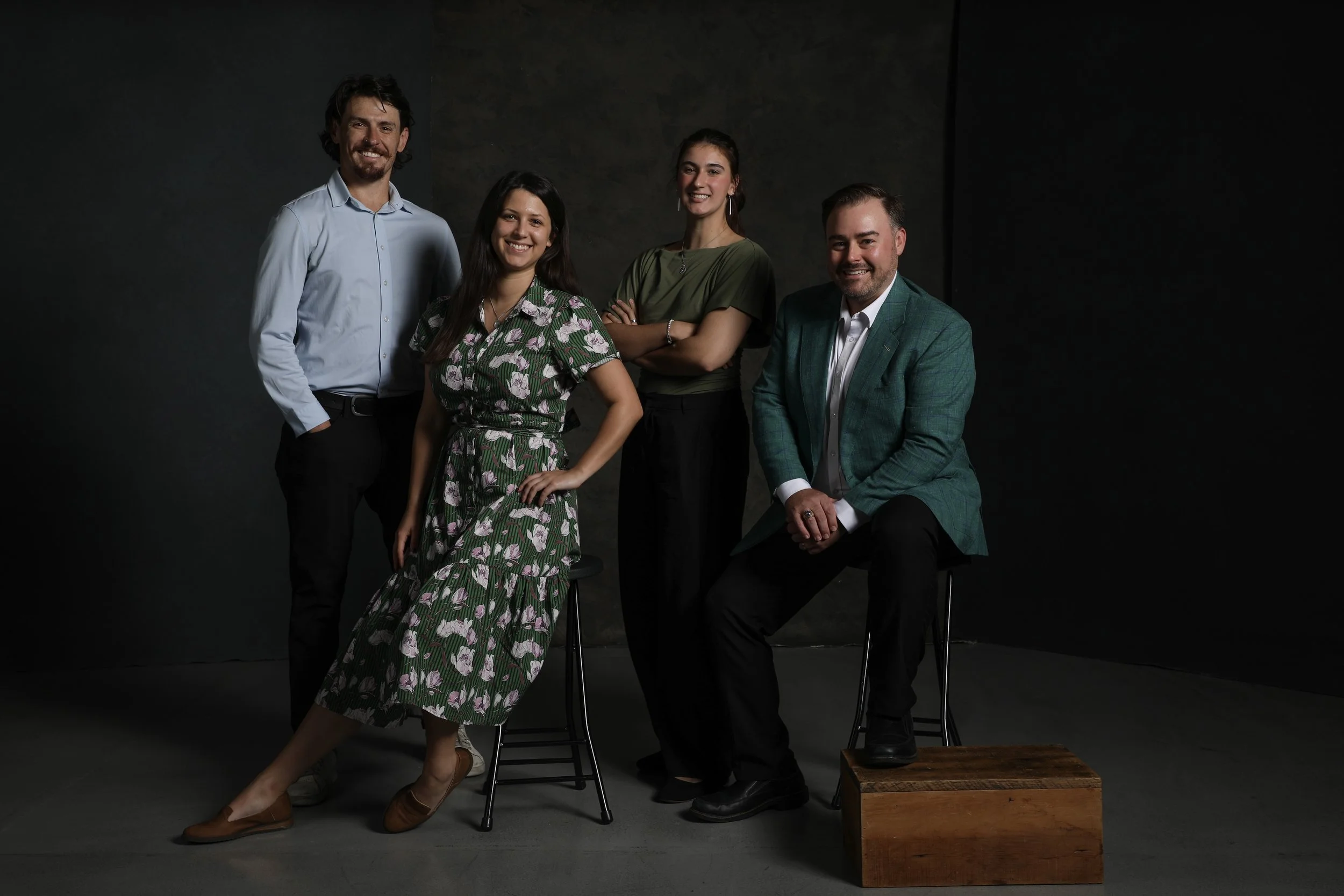 A group of five people, three men and two women, posing together in a studio with a dark background. They are smiling and dressed in semi-formal attire, with one woman in a floral dress, and the others in shirts and jackets.