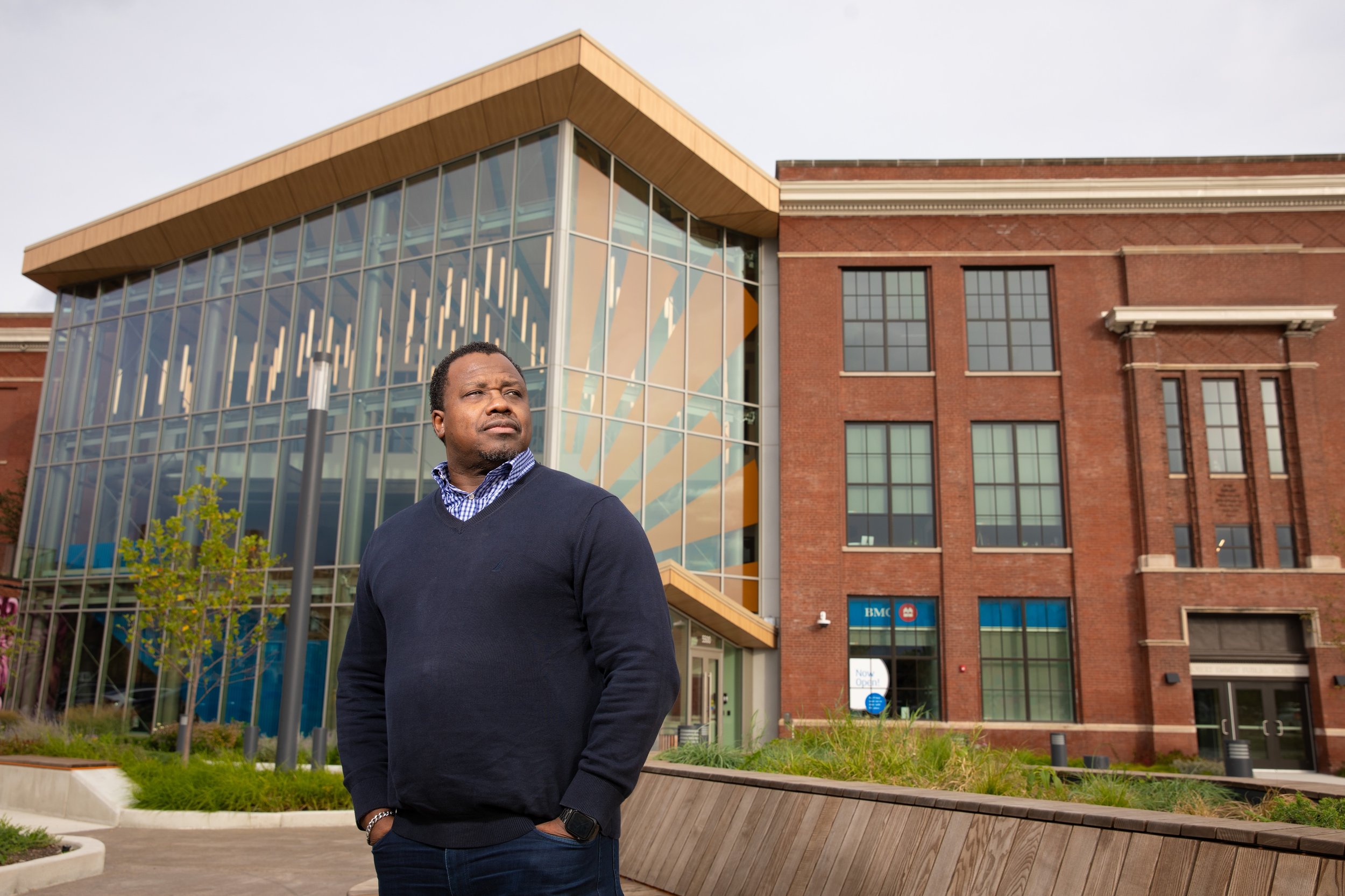 A man standing outside in front of a modern brick and glass building, looking off to the side with a serious expression.