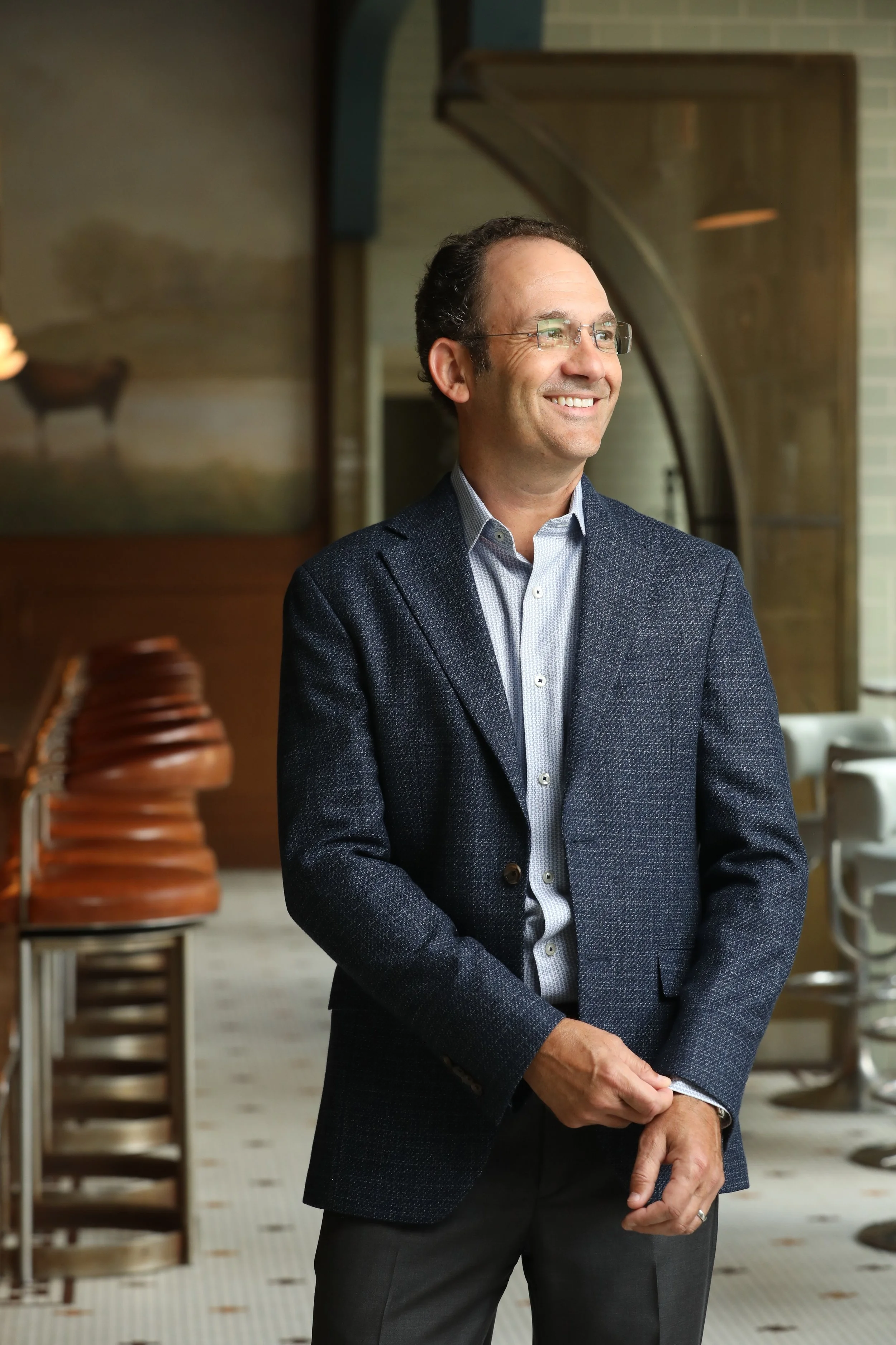 A man in a dark blue blazer and glasses smiling, standing indoors with a row of brown chairs and a decorative wall in the background.