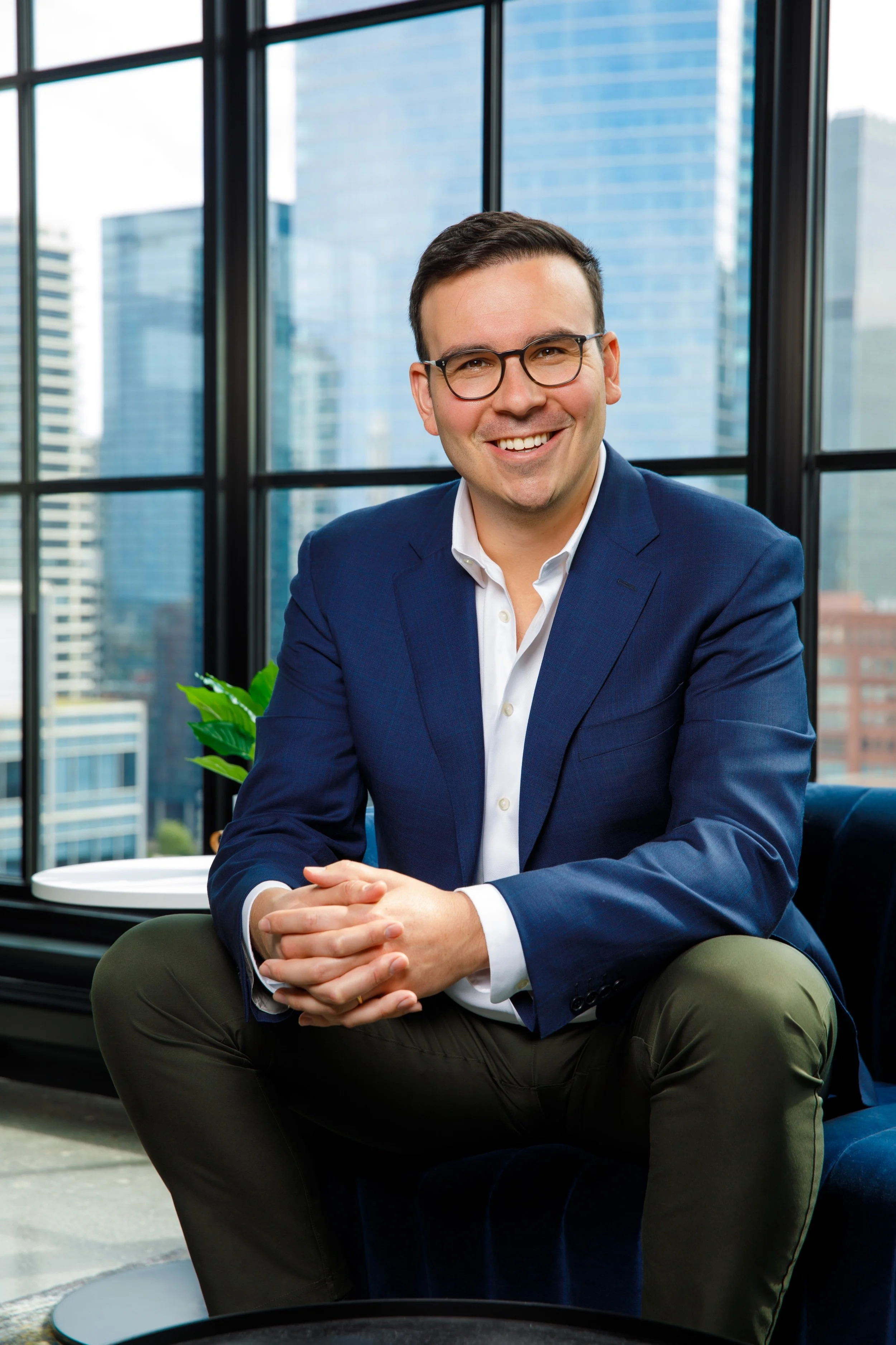 A smiling man in a navy blazer and white shirt, wearing glasses, sitting in a modern office with large glass windows showing city skyscrapers.