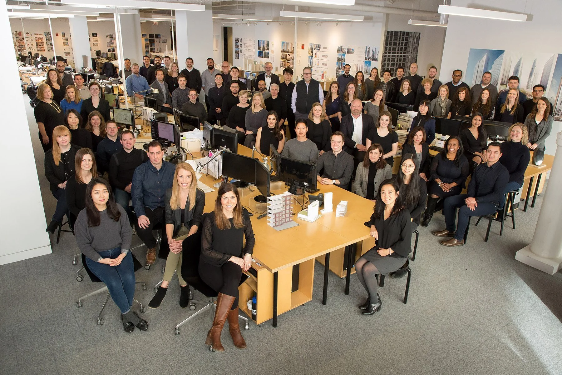 A large group of diverse professionals gathered in a modern office space, posing for a group photo around a central table with multiple computer monitors and architectural models.