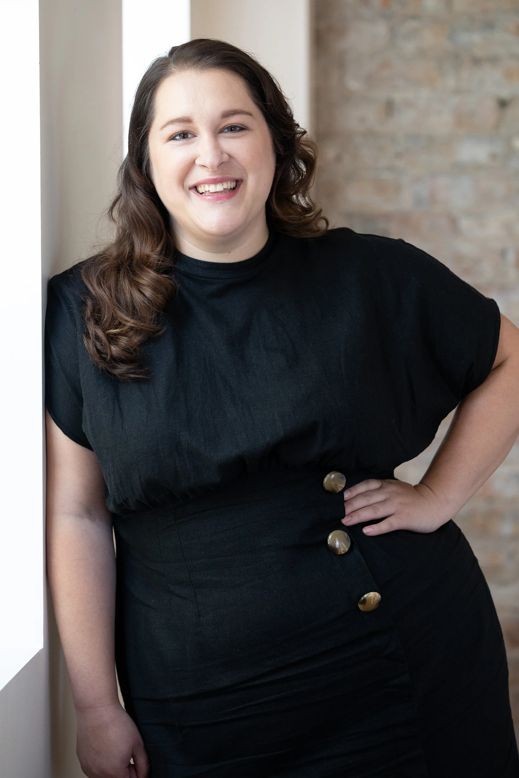 A woman with long, wavy brown hair, smiling, wearing a black top with large buttons, standing indoors near a window and brick wall.