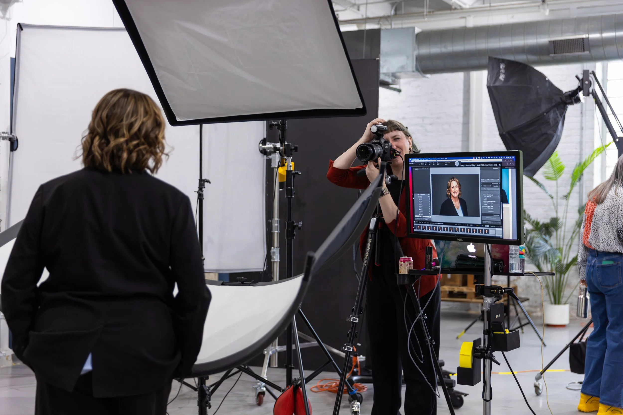 A woman with short hair taking a portrait photograph of a smiling woman on a computer monitor during a photo shoot in a studio.
