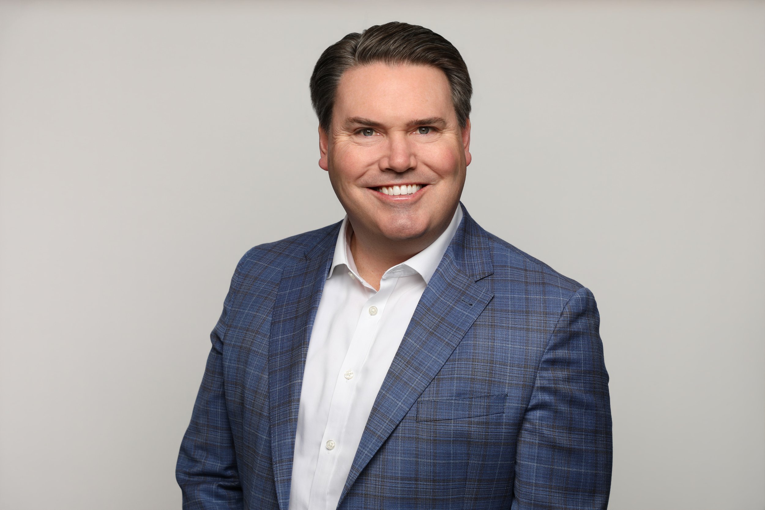 A smiling man with dark hair, wearing a blue checked blazer and a white dress shirt, posing against a plain light background.