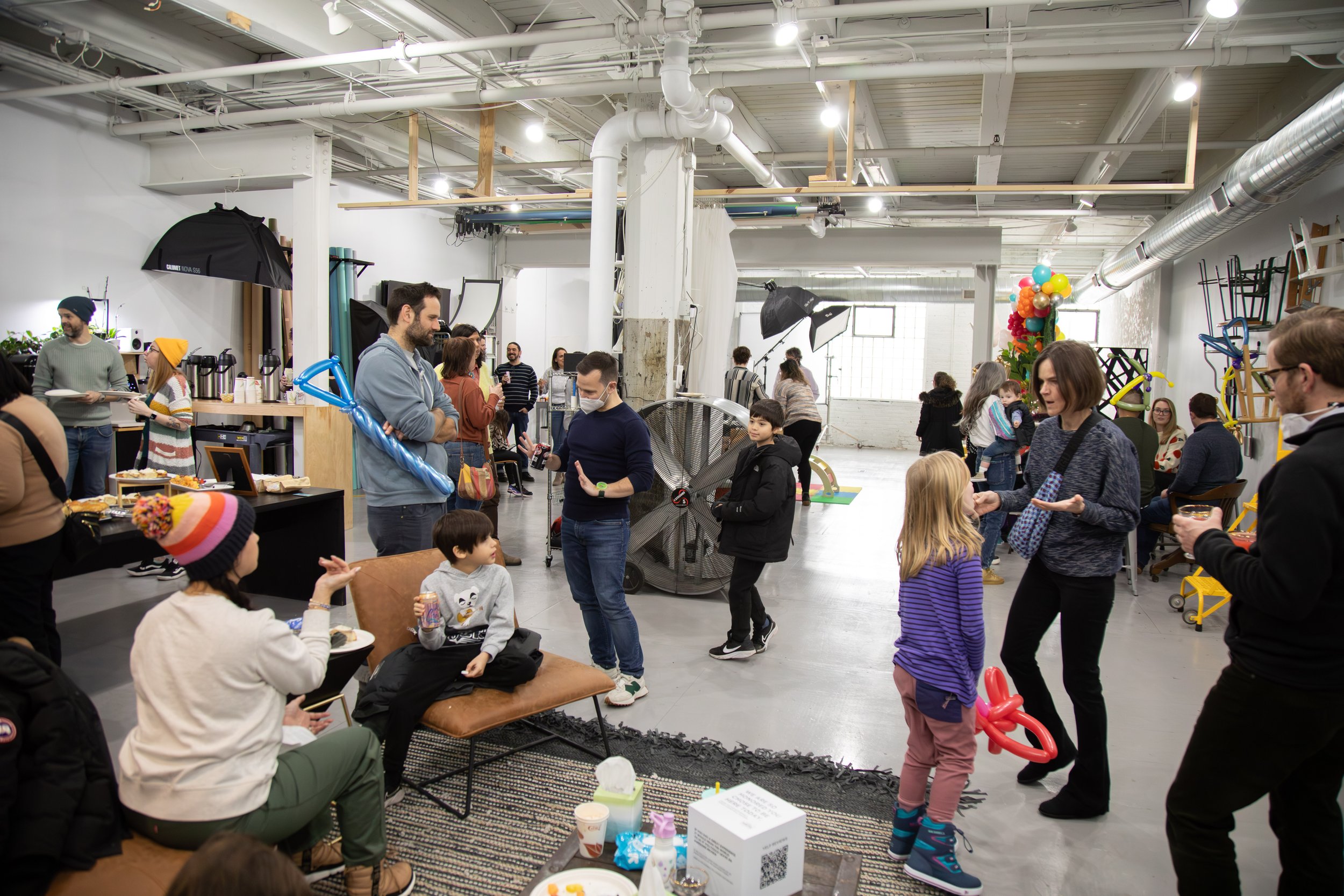 People gathered in a large, industrial-style indoor space with white walls, high ceilings, and exposed pipes. Some individuals are sitting and eating, while others stand and converse. There are balloons, children playing, and photography equipment se