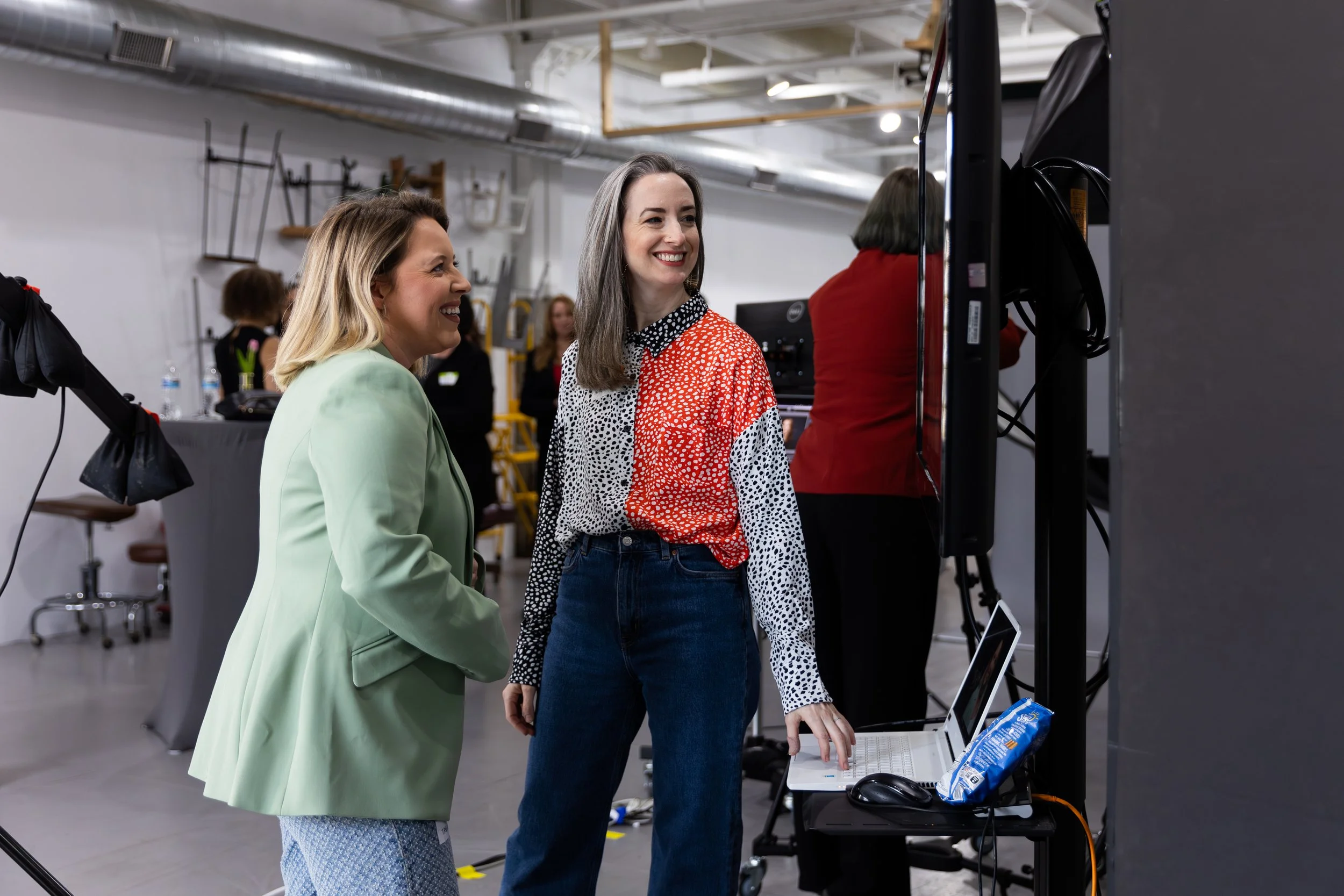 Two women smiling and interacting with a computer at an event or workspace. One woman wears a light green jacket and the other a red and white patterned shirt.
