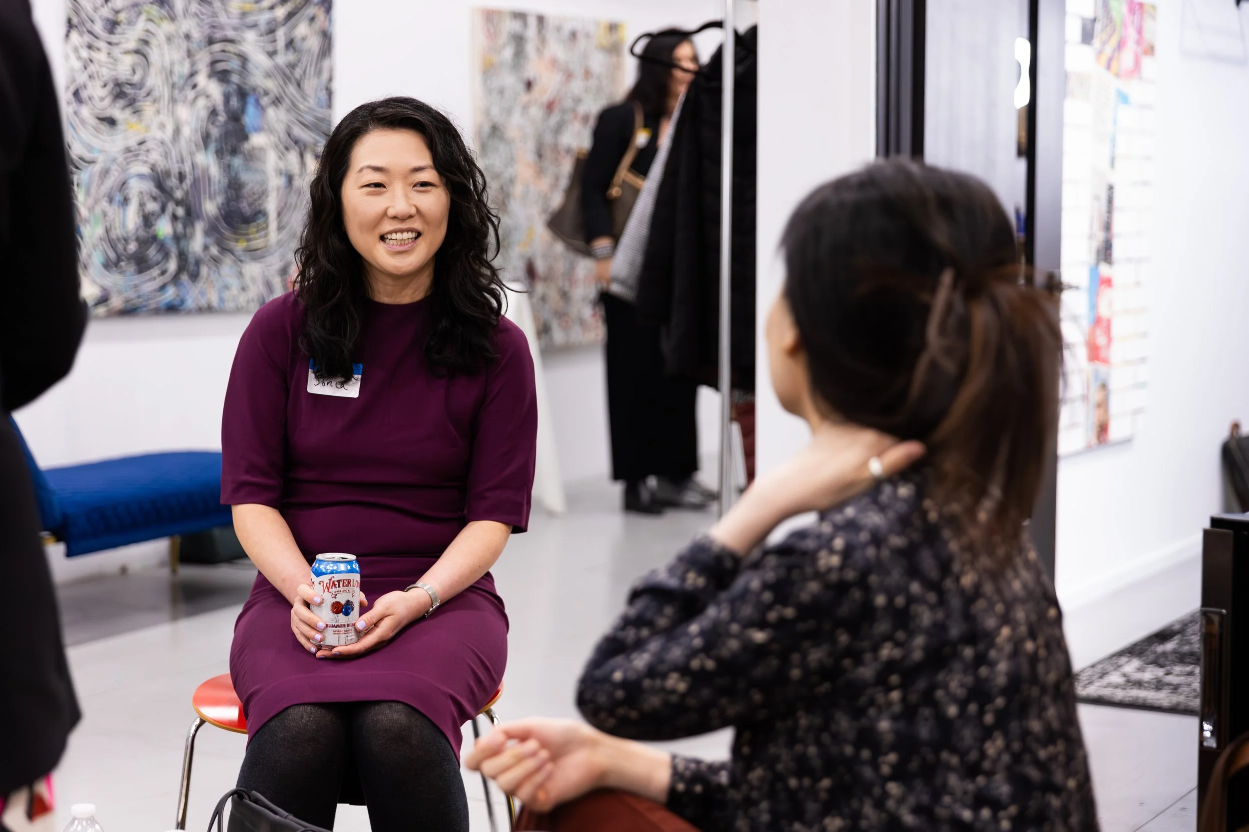 A woman with long curly black hair wearing a purple dress smiling while sitting on a small chair during an indoor event, holding a can of Waterloo sparkling water. Two other women are partially visible, one facing away and the other in the background