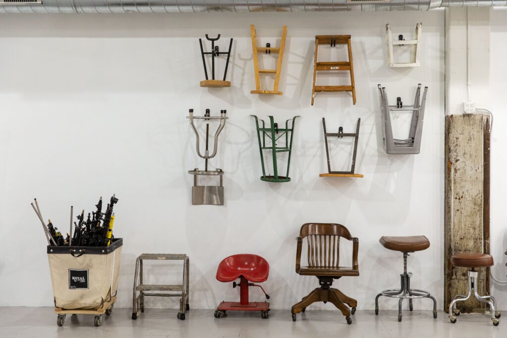 Collection of chairs hanging on a white wall, various types of chairs and stools below, with a cart filled with tools on the left, and a long vertical piece of wood on the right.