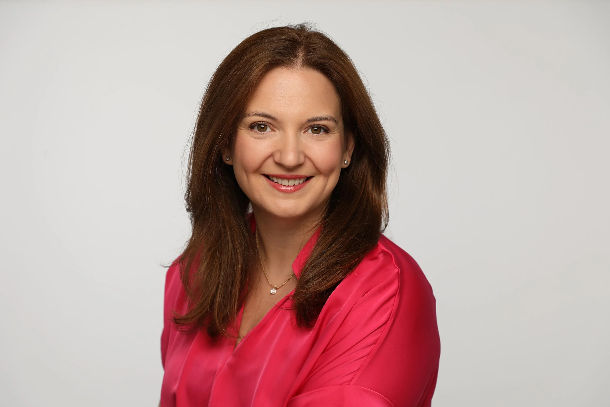 A woman with long brown hair wearing a pink top and jewelry, smiling in front of a plain light gray background.