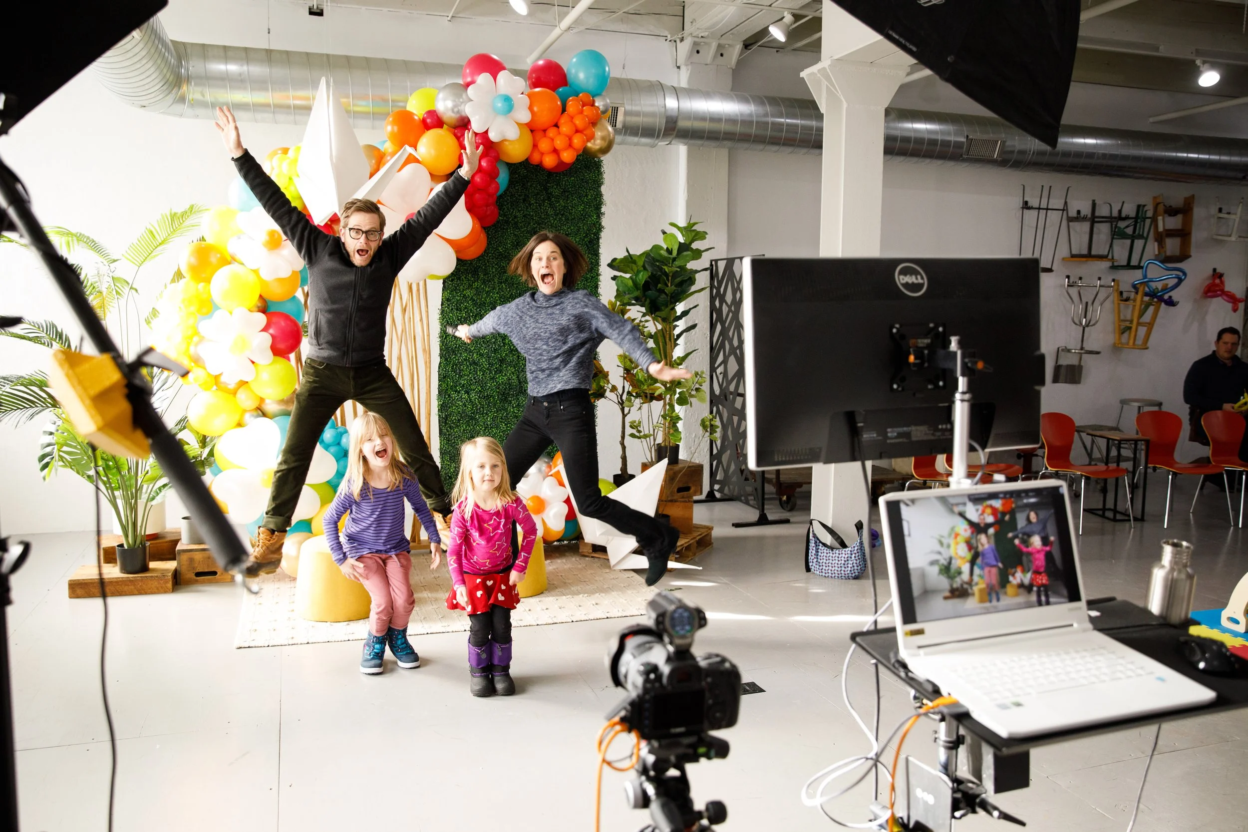 Group of children and adults celebrating behind the scenes of a photo shoot with balloon decorations, camera equipment, and a backdrop.