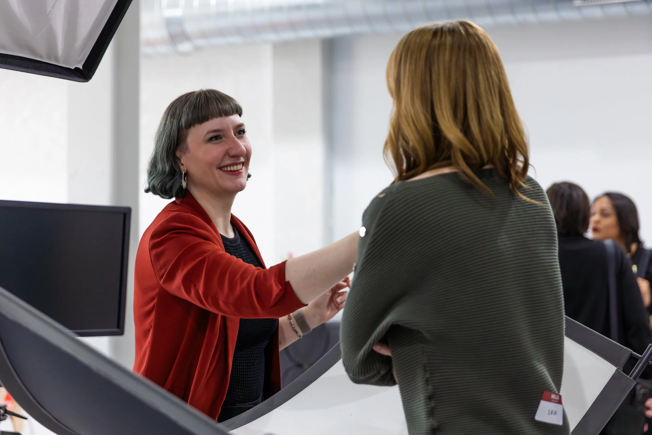 Two women exchanging a handshake at a reception or conference, seen through the reflection of a mirror or display on a piece of electronic equipment.