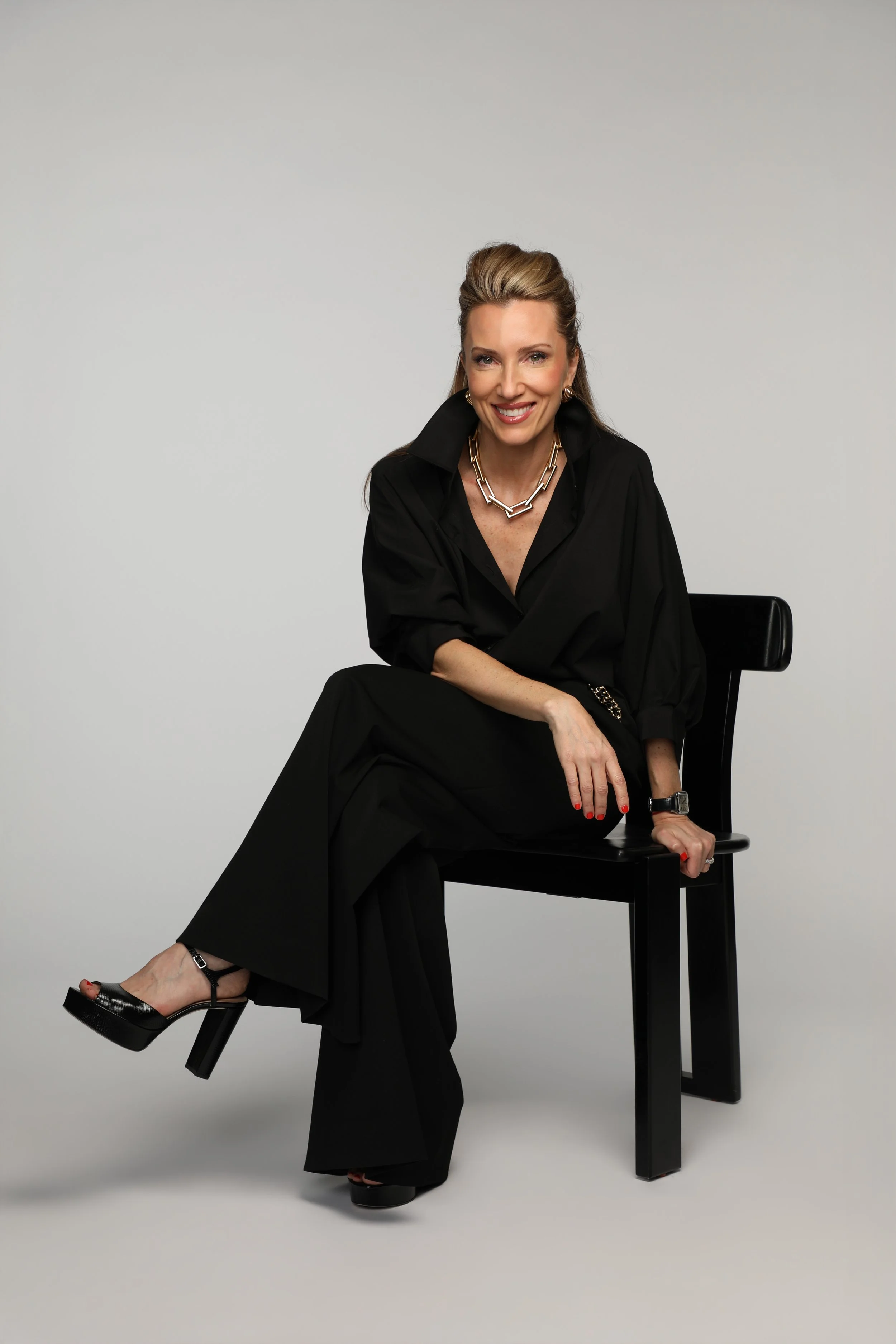 A woman sitting on a black chair against a plain light gray background, smiling, dressed in black with jewelry, with one leg crossed over the other, wearing high-heeled platform shoes.