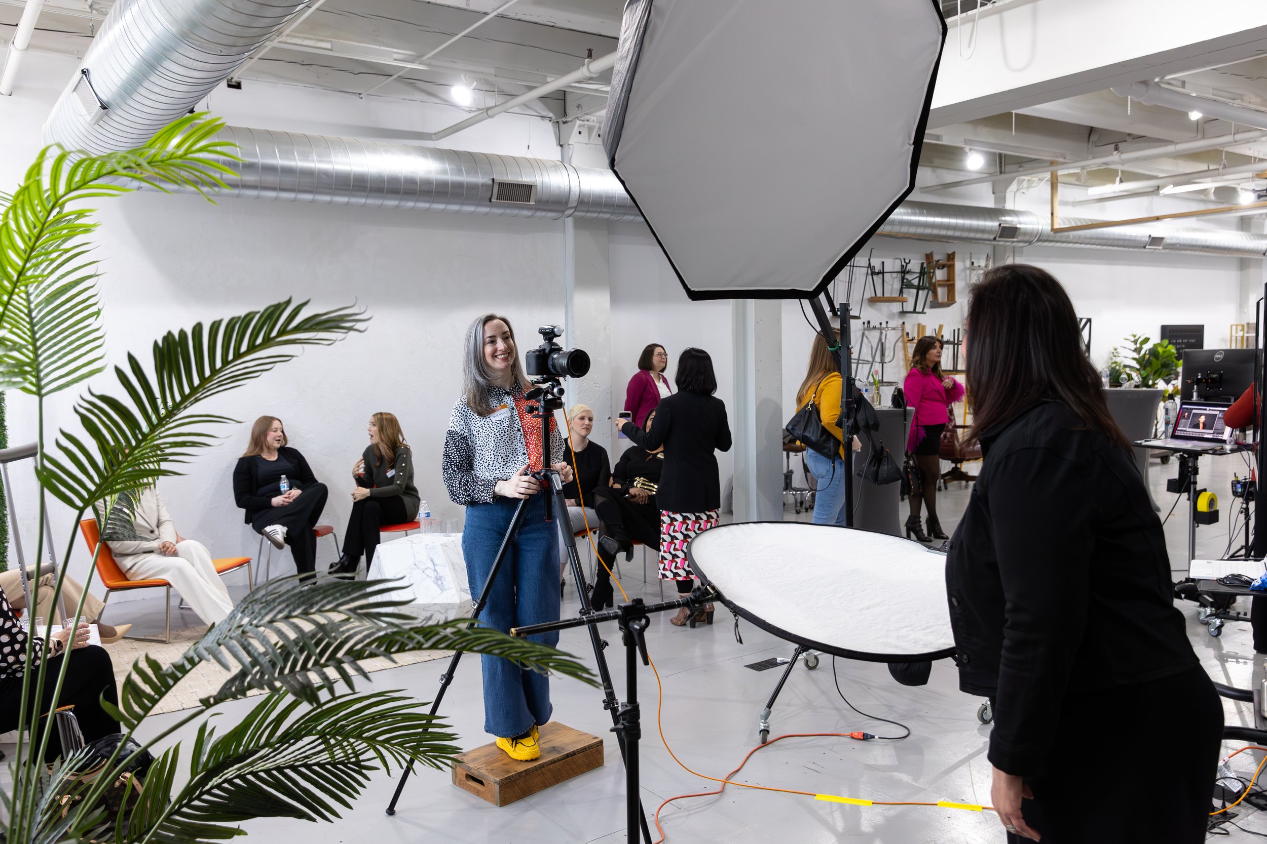 A woman in yellow shoes and blue jeans standing on a wooden box, smiling at a camera in a photography studio. Several people are seated and standing in the background, with a large softbox light overhead and plants in the foreground.