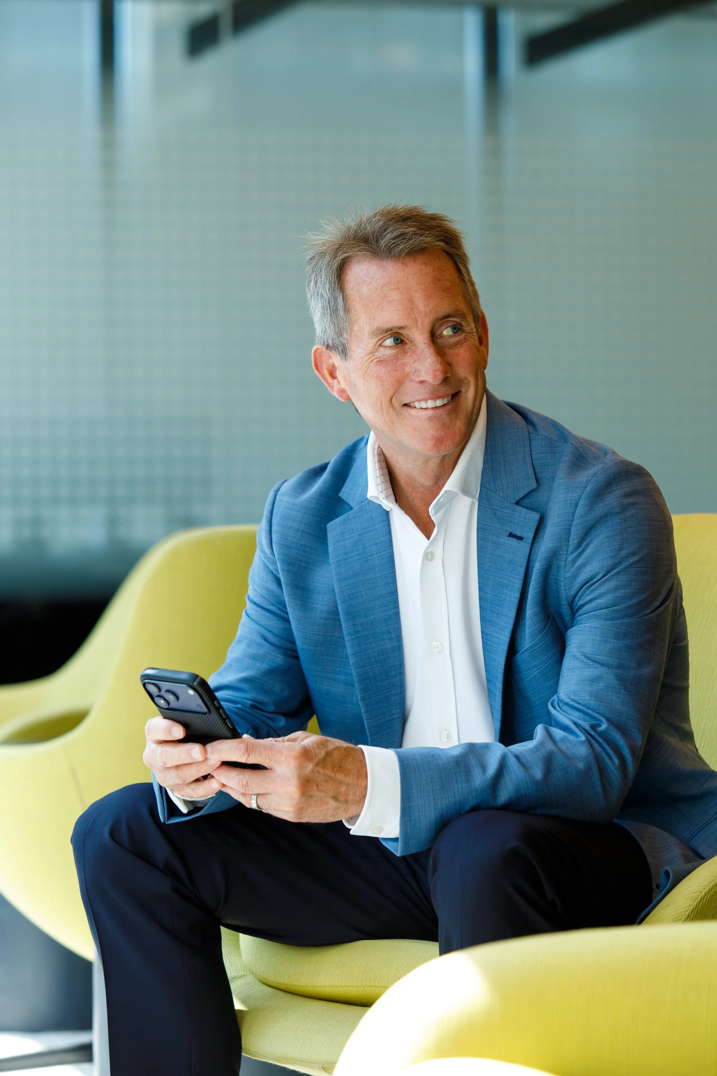 A professional man in a blue blazer sitting on a yellow chair looking up from his phone