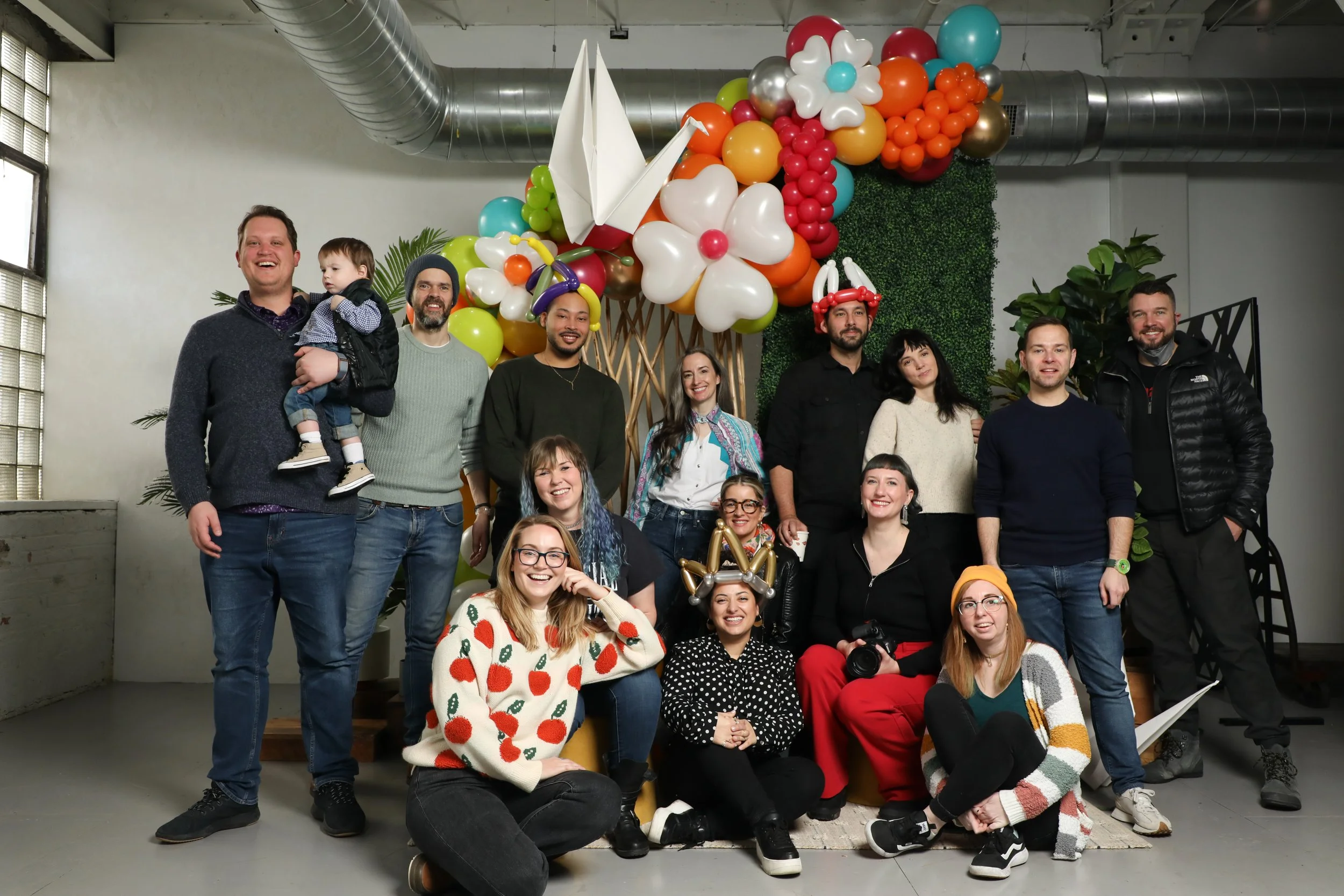 Group of people smiling and posing together in front of a colorful balloon display, indoors with industrial-style ceiling vents and greenery in the background.