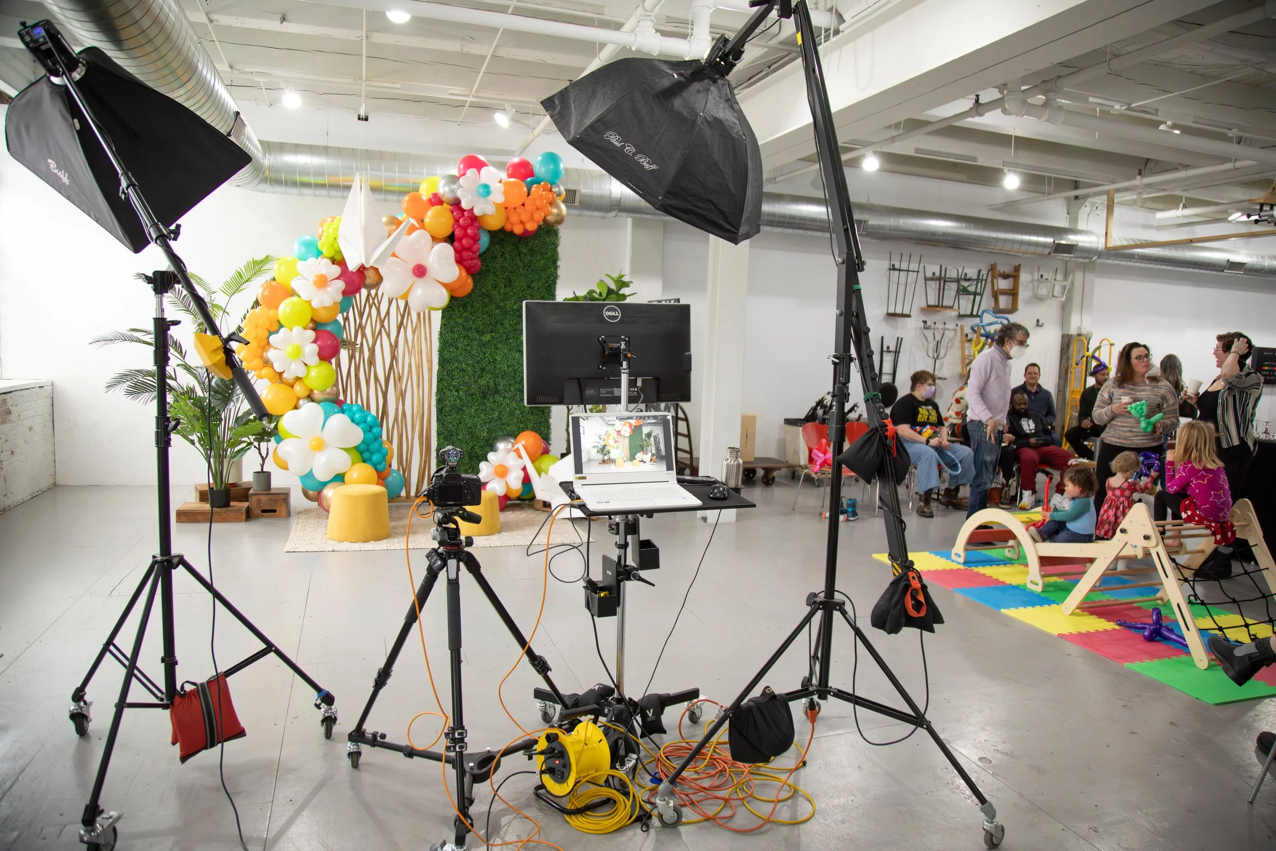 Photography studio setup with professional lighting equipment and a backdrop decorated with colorful balloons and flowers for a children's event, with people including children and adults gathered in the background.