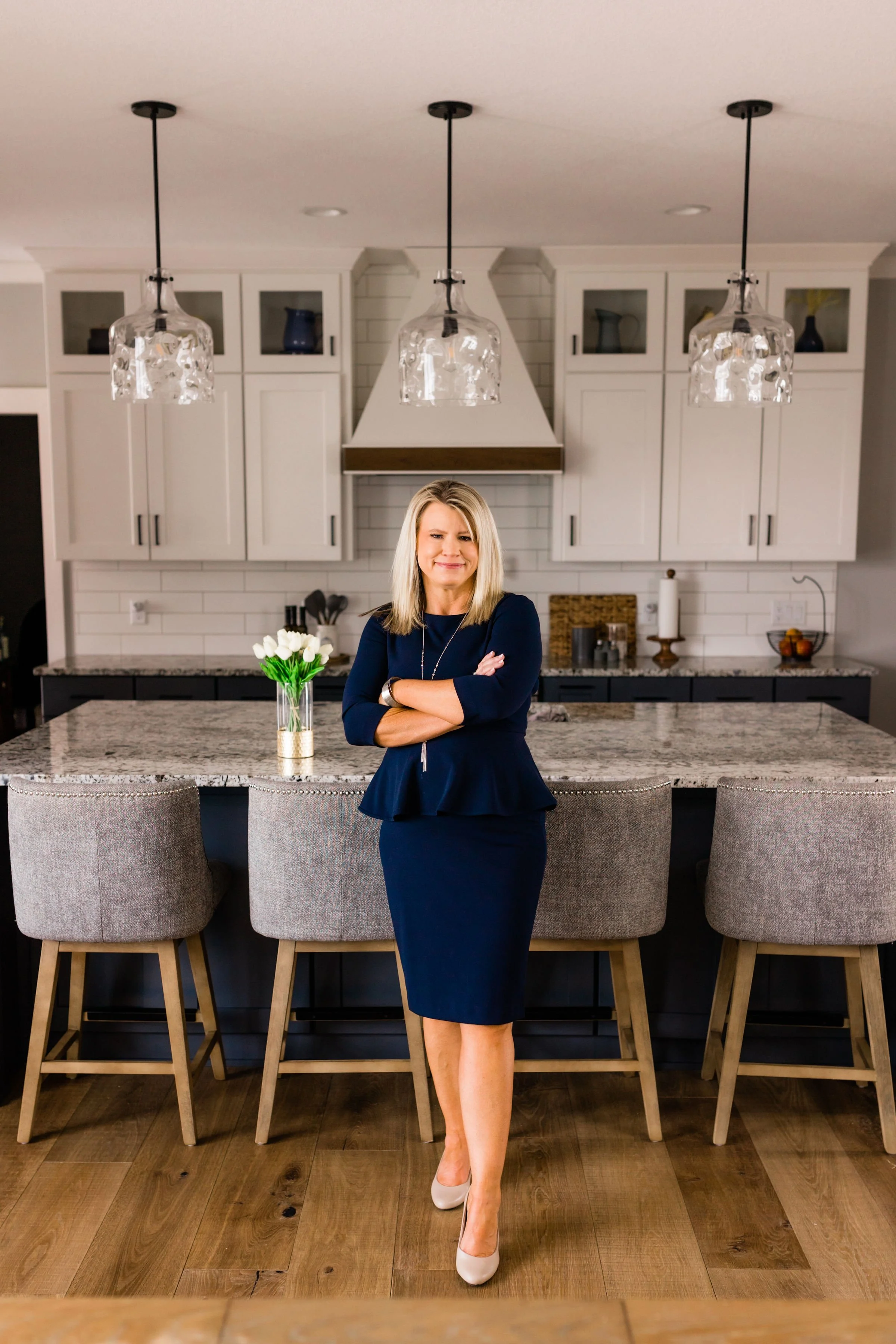 Woman in business attire standing in modern kitchen with marble countertops, gray bar stools, pendant lights, and white cabinetry.