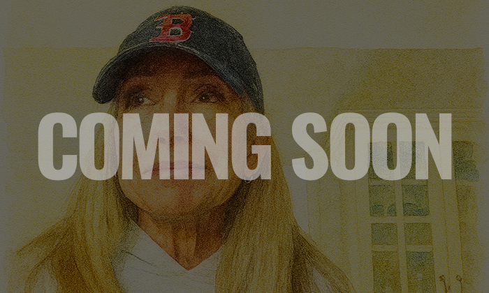 A woman wearing a dark baseball cap with a red B on it, standing indoors with a beige wall and a window behind her. The words "COMING SOON" are overlaid in large white letters.