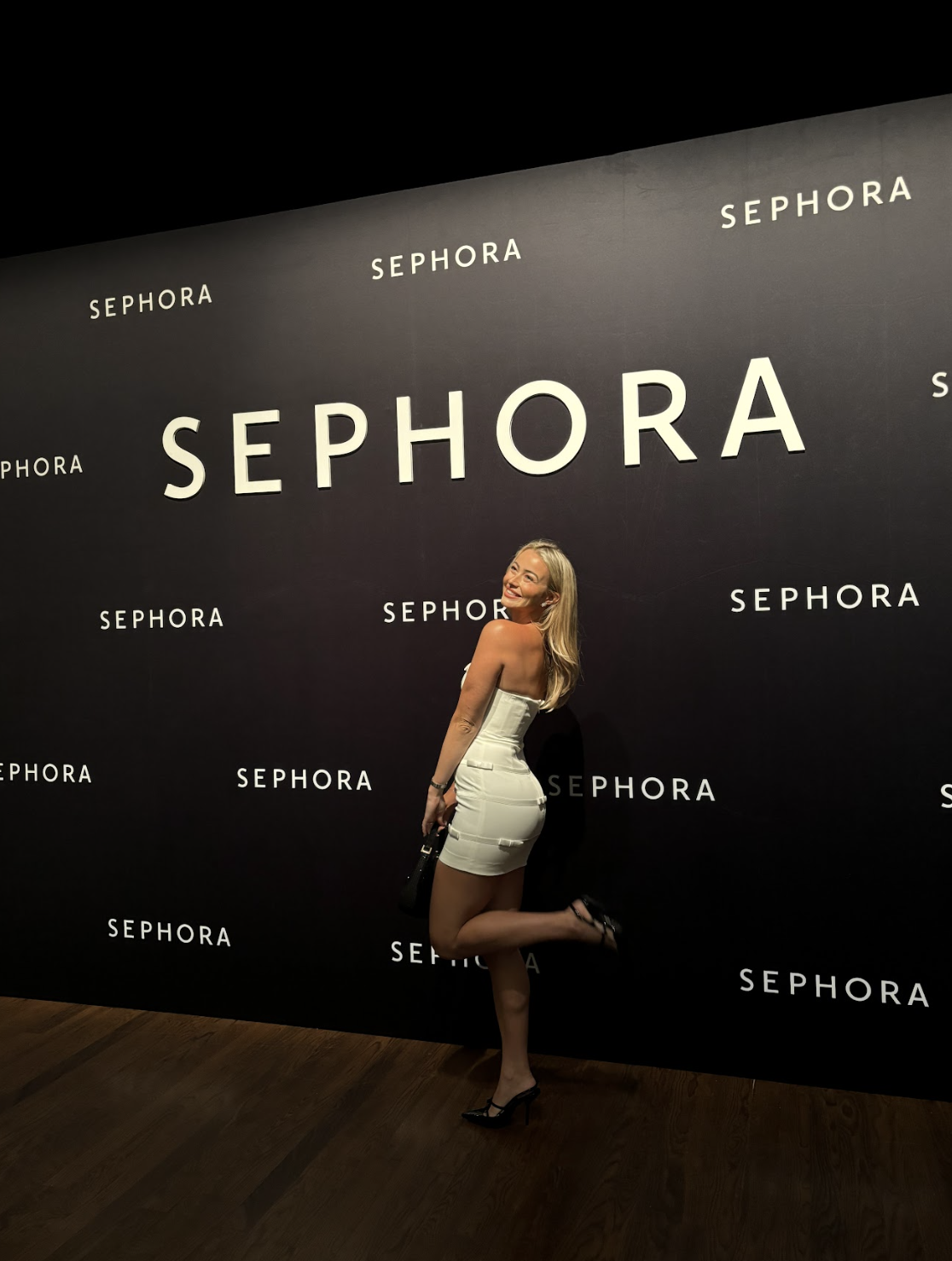 A woman in a white dress posing against a black Sephora branded wall, smiling and lifting one leg.