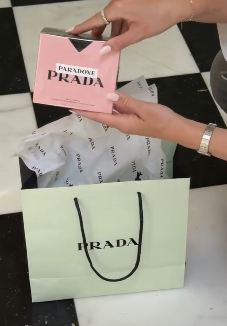 A person is holding a pink Prada box with the word 'Paradoxe' on it, above a shopping bag with the Prada logo, on a black and white checkered floor.