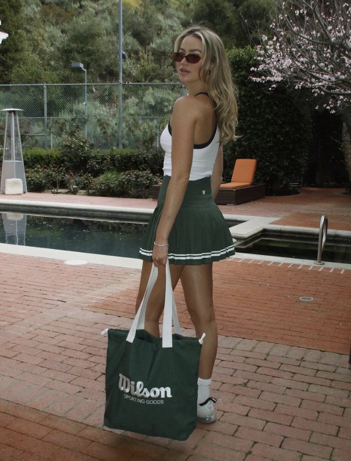 A woman wearing sunglasses, a white tank top, and a dark green tennis skirt standing on a brick patio near a swimming pool, holding a Wilson sporting goods bag.