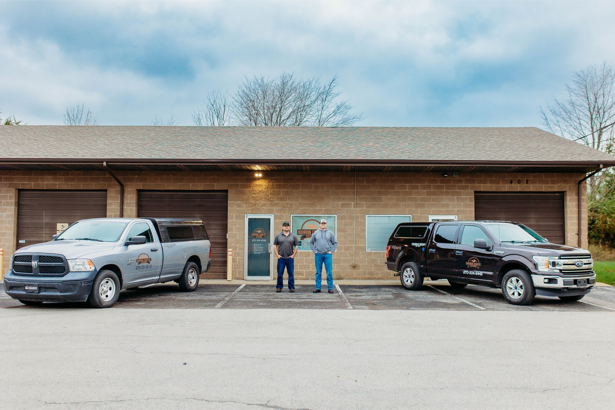 Two men standing in front of a building with two parked trucks, each displaying a business logo, in an outdoor setting.