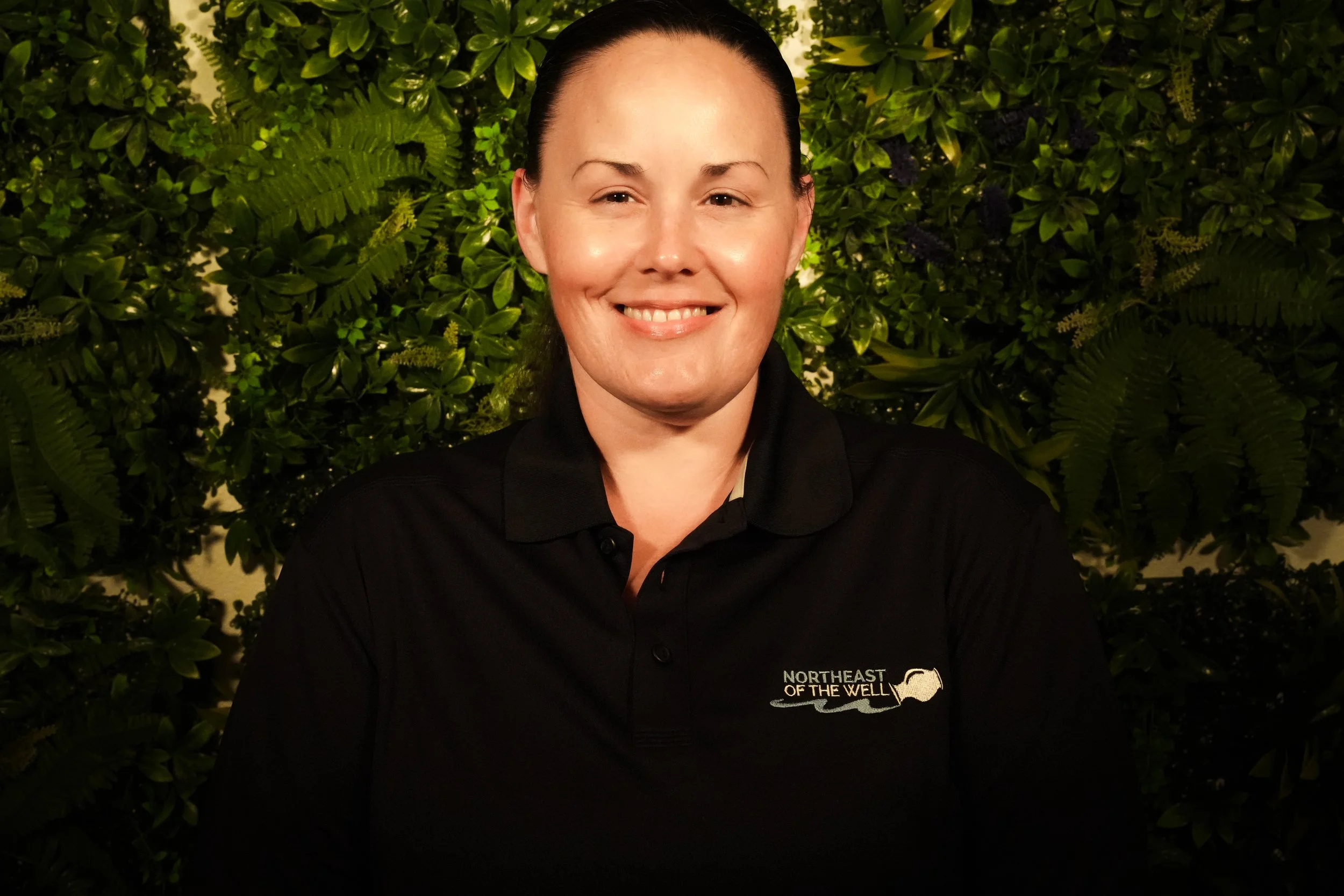 A woman with dark hair tied up, wearing a black top with a floral patterned sheer cover-up, smiling in front of a green leafy background.