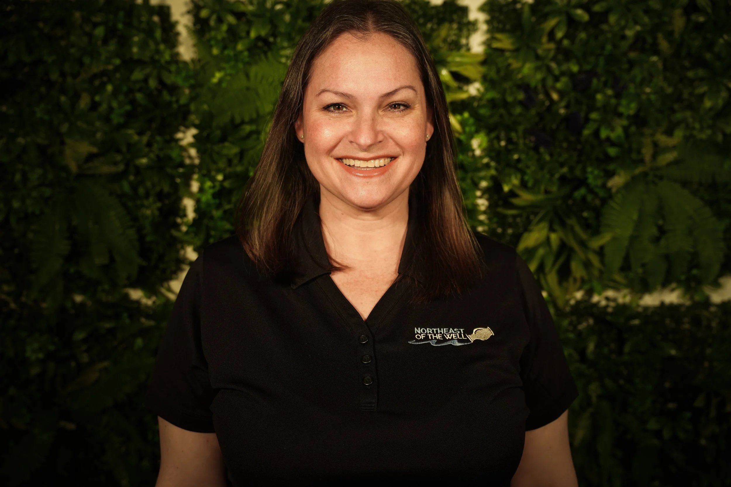 A woman with long brown hair wearing a black top with lace detail, smiling in front of a green leafy background.