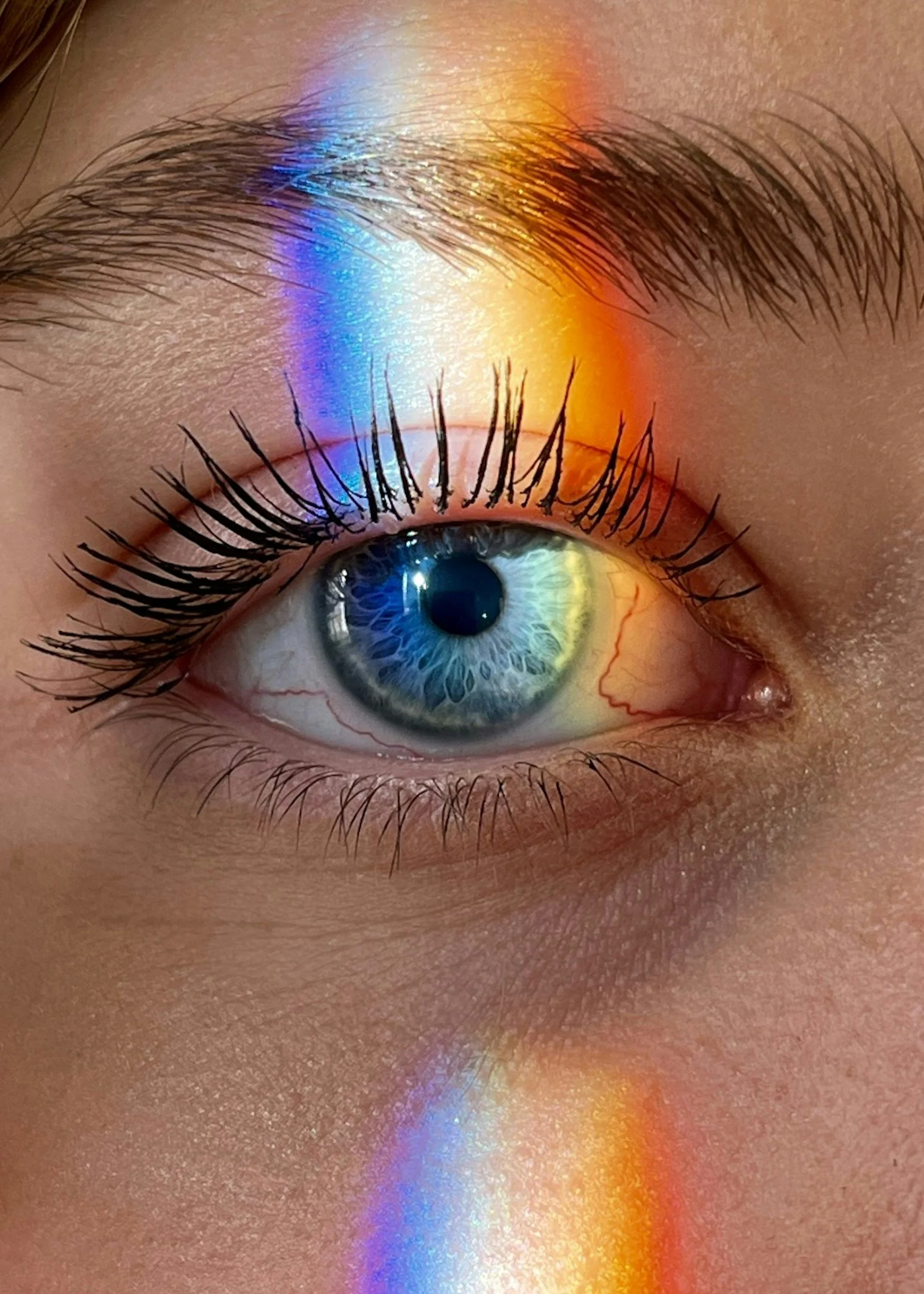 Close-up of a human eye with a rainbow-colored light and reflection on the eyelid and surrounding skin.