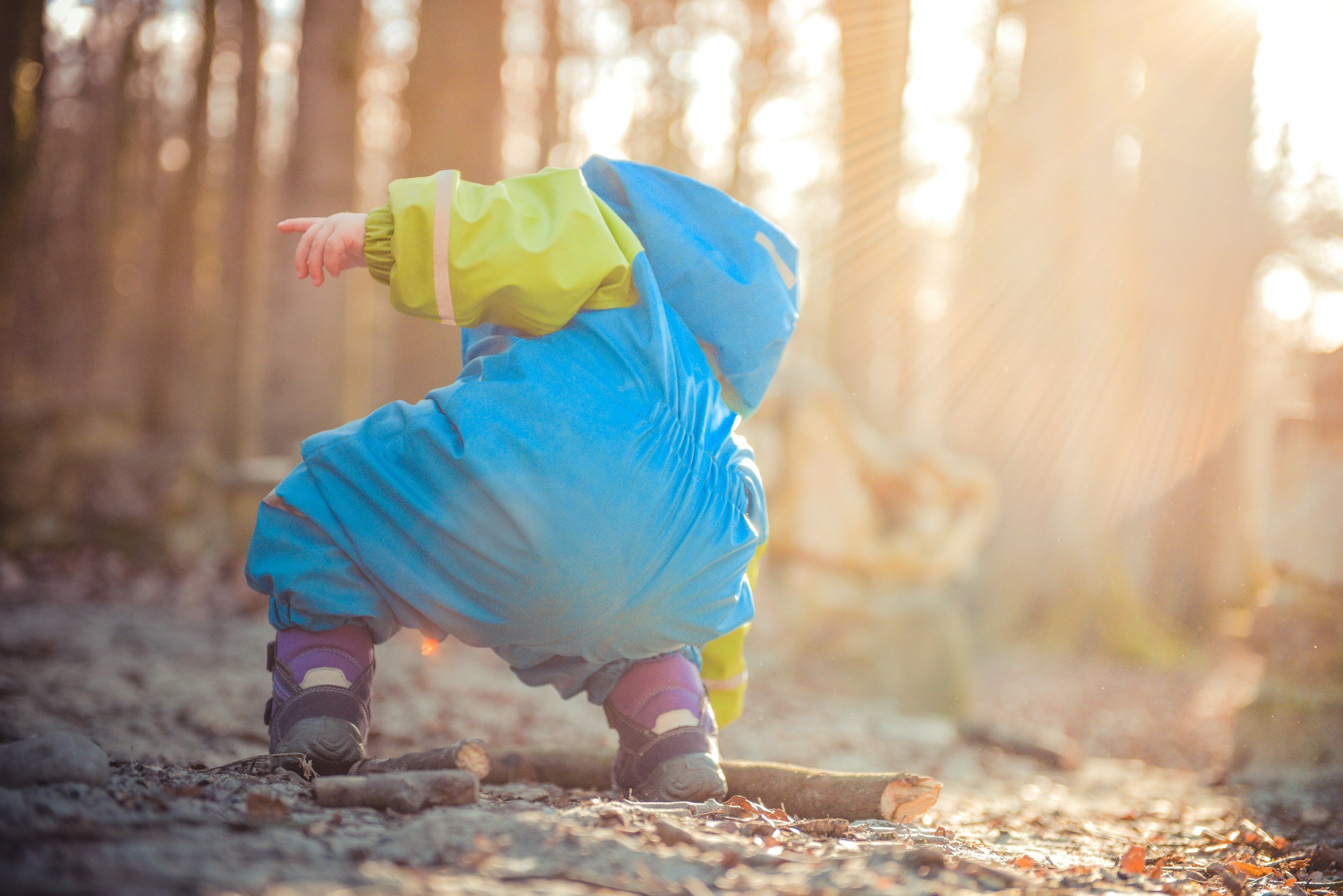 A young child wearing a green and blue jacket, gray pants, and pink shoes is crouching in a wooded area during autumn with fallen leaves on the ground, illuminated by warm sunlight.