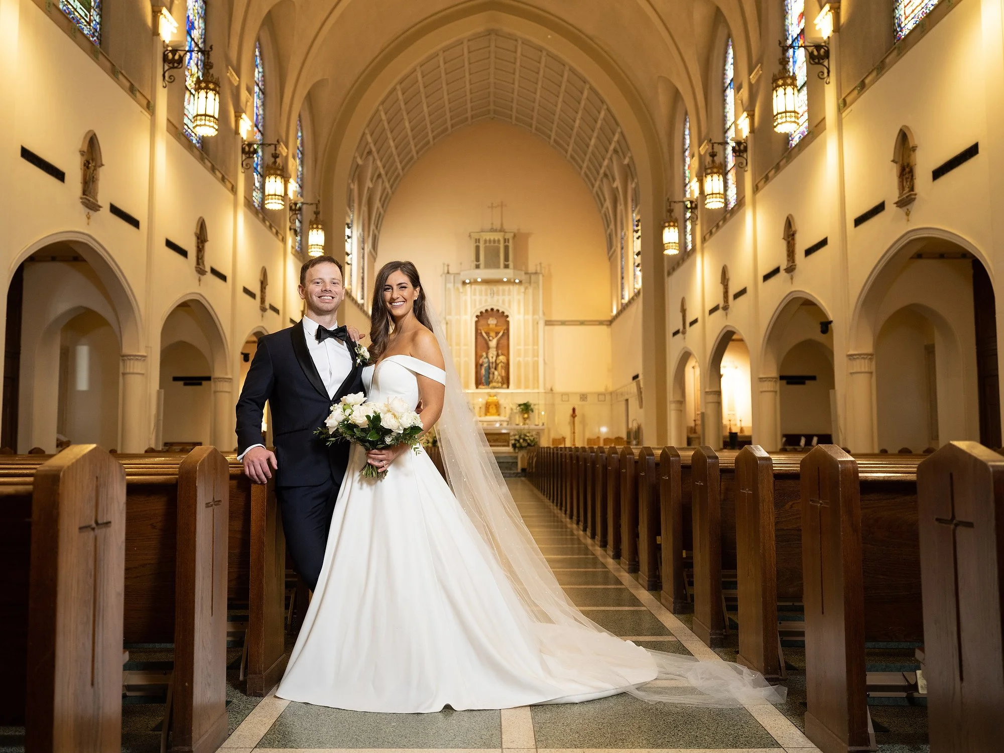 A newlywed couple standing inside a church, smiling at the camera. The bride is holding a bouquet of white flowers and wearing an off-the-shoulder wedding gown with a long veil. The groom is dressed in a black tuxedo with a bow tie. The church interi