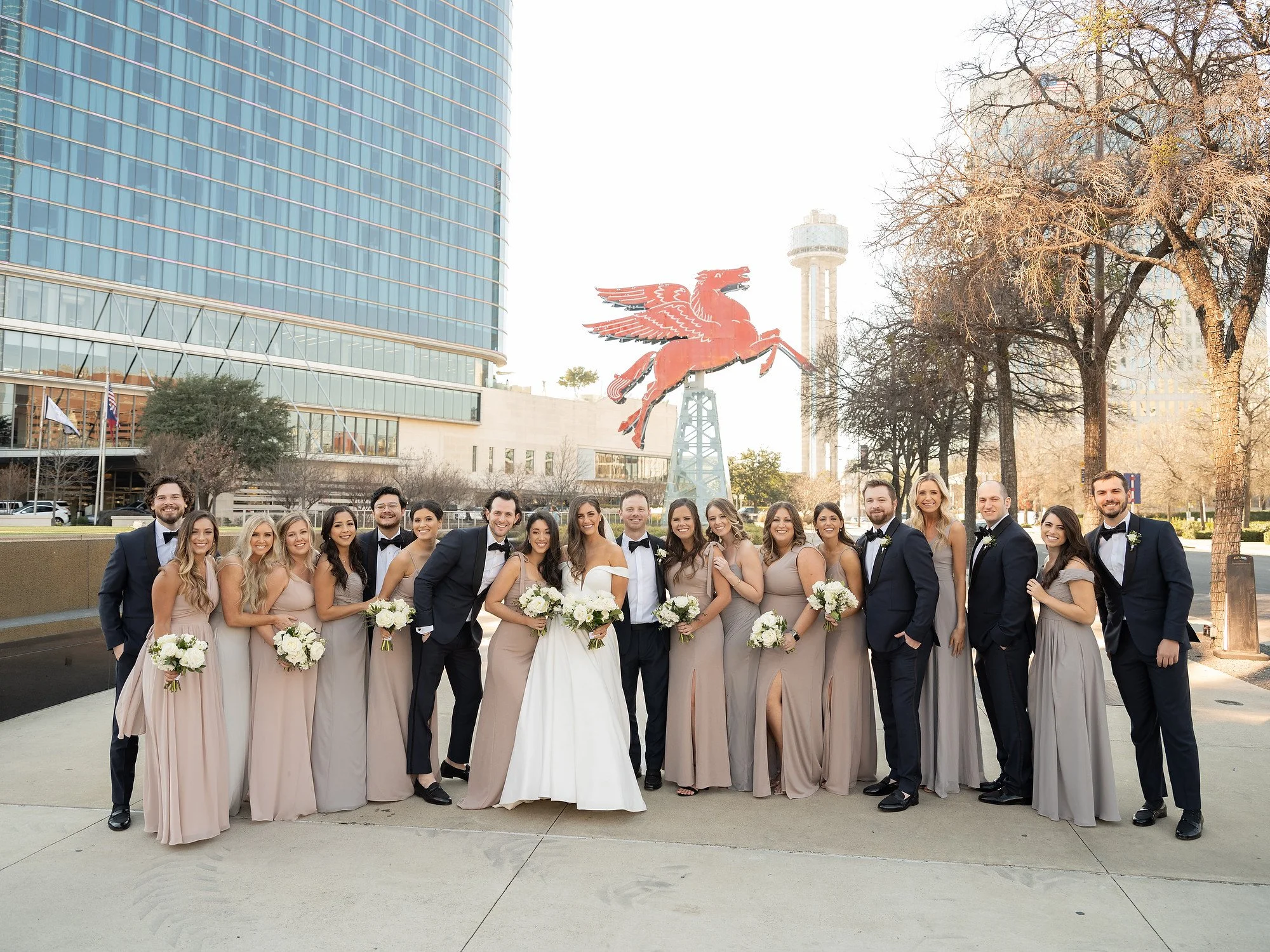 Group of wedding party members, including bridesmaids in beige dresses and groomsmen in dark suits, standing outdoors in front of the Las Vegas Strip with the Welcome to Fabulous Las Vegas sign and the Stratosphere Tower visible in the background.