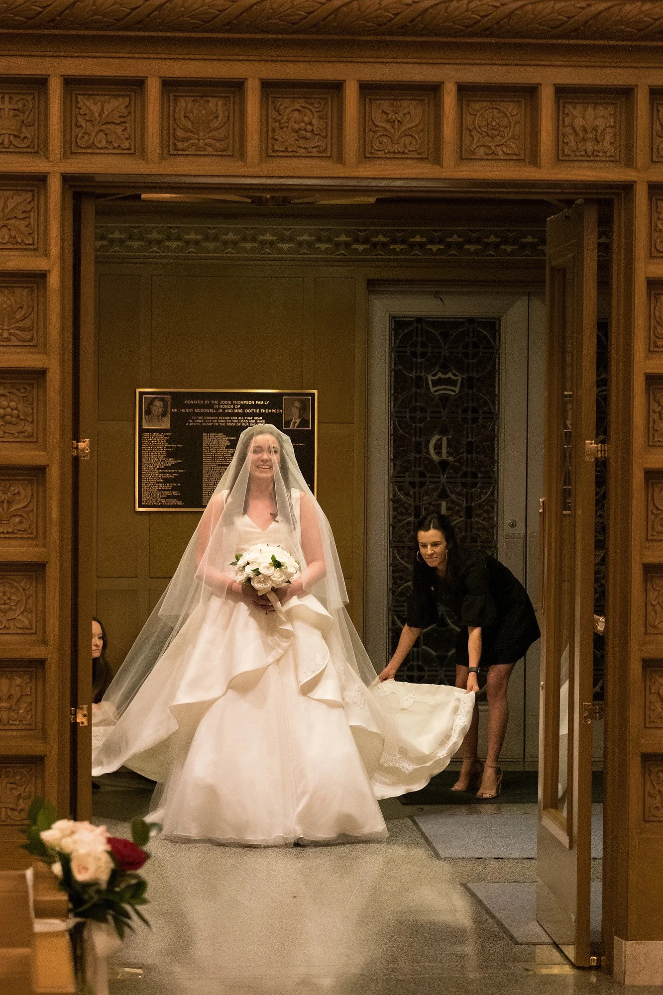 Bride in a wedding dress holding a bouquet, smiling inside a church with two women helping her with her dress.