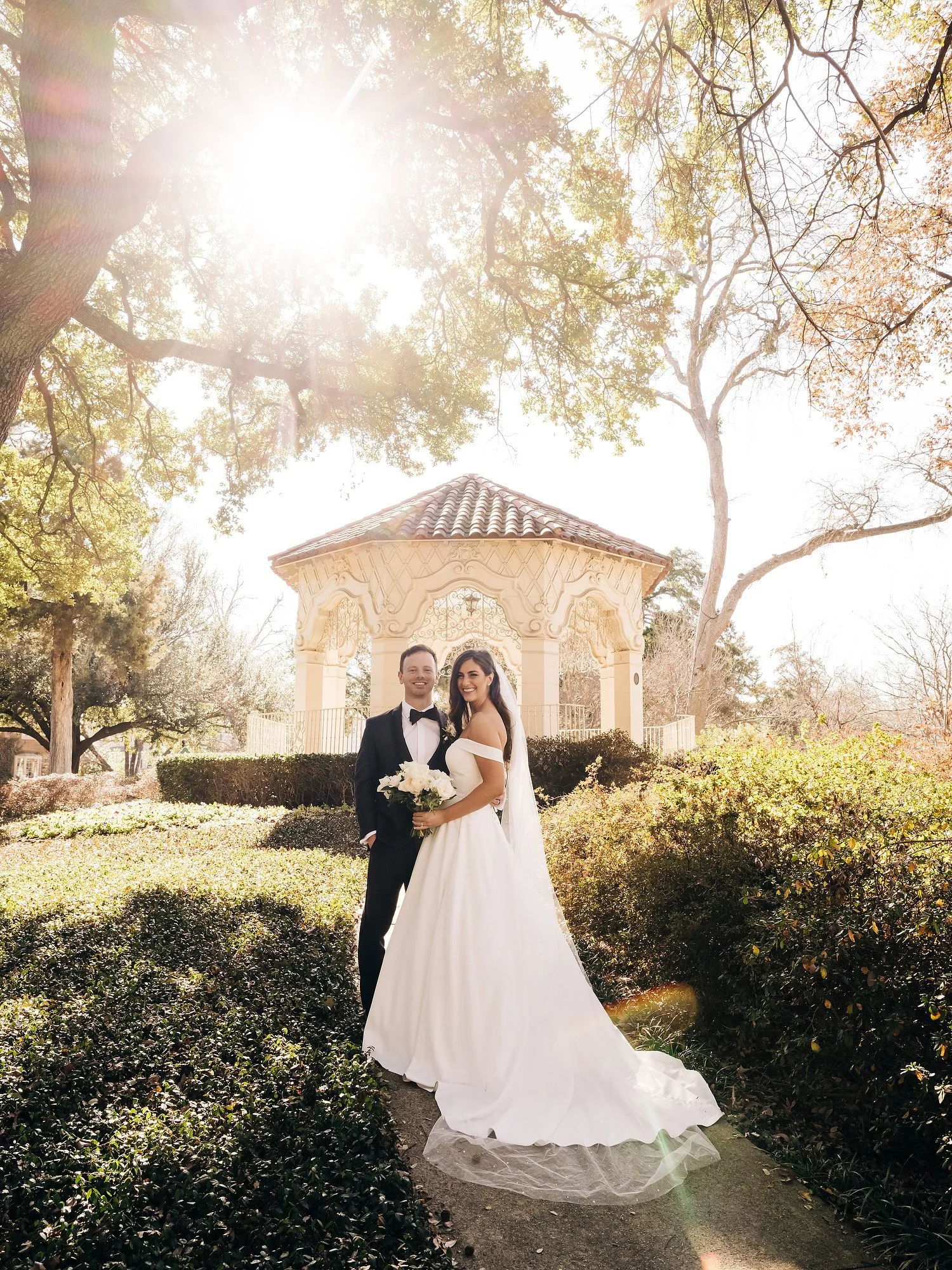 A bride and groom in wedding attire standing on a garden path, smiling, with a gazebo and trees in the background, in bright sunlight.