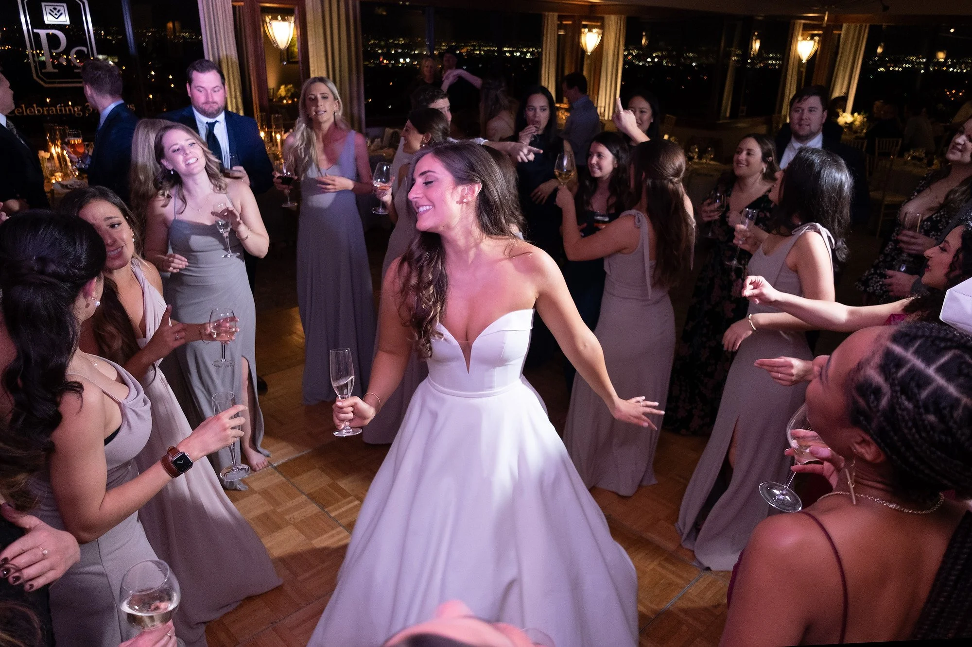 A bride in a white wedding gown dancing and smiling on a dance floor surrounded by wedding guests holding glasses of champagne, celebrating at a wedding reception in a decorated indoor venue with large windows and city lights outside.