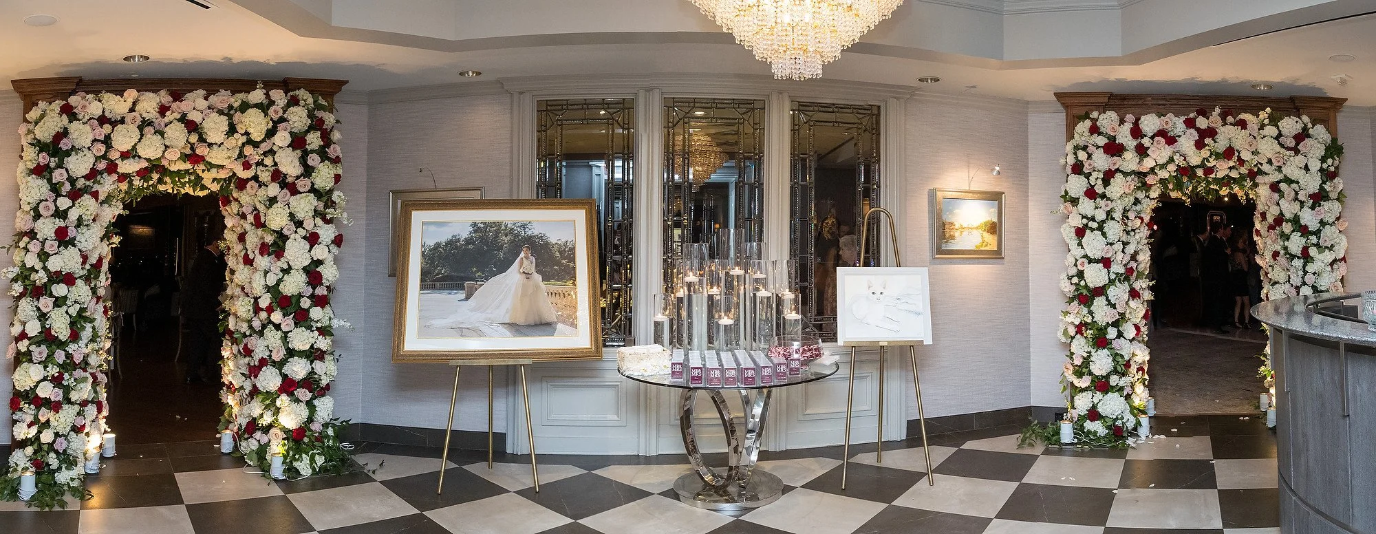 Elegant wedding reception entrance with two large floral arches made of white, pink, and red flowers, a framed wedding photograph, artwork of a white cat on an easel, and a checkered black and white floor.