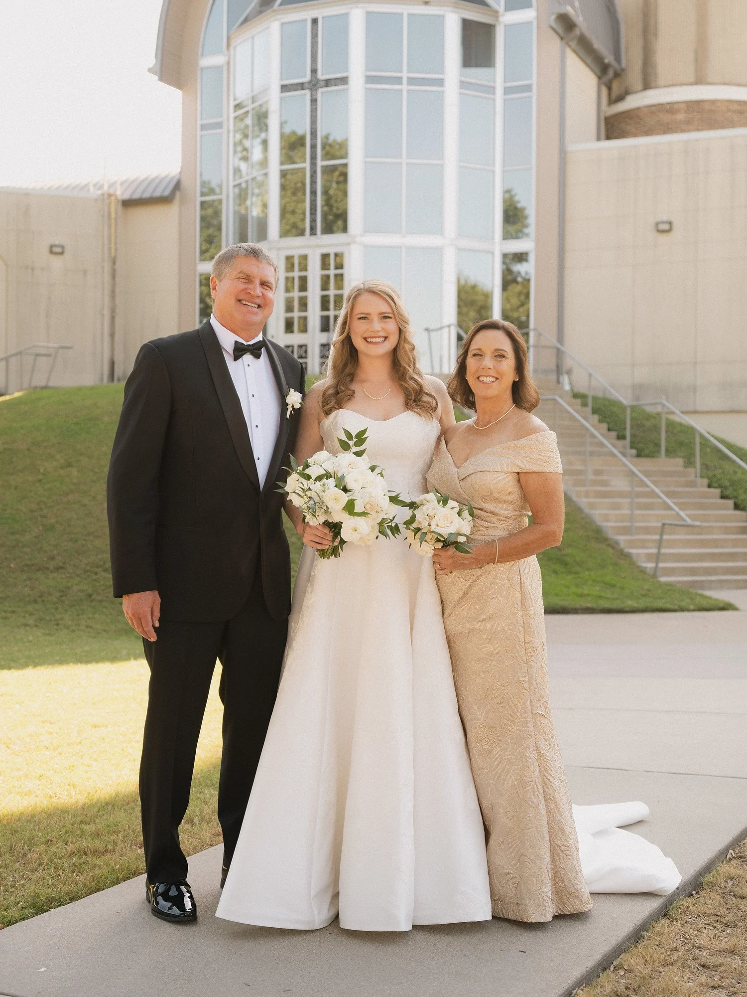 A bride in a white wedding gown standing between her father in a black tuxedo and her mother in a gold dress, all smiling, holding bouquets of white flowers, outside in front of a modern building with large glass windows.
