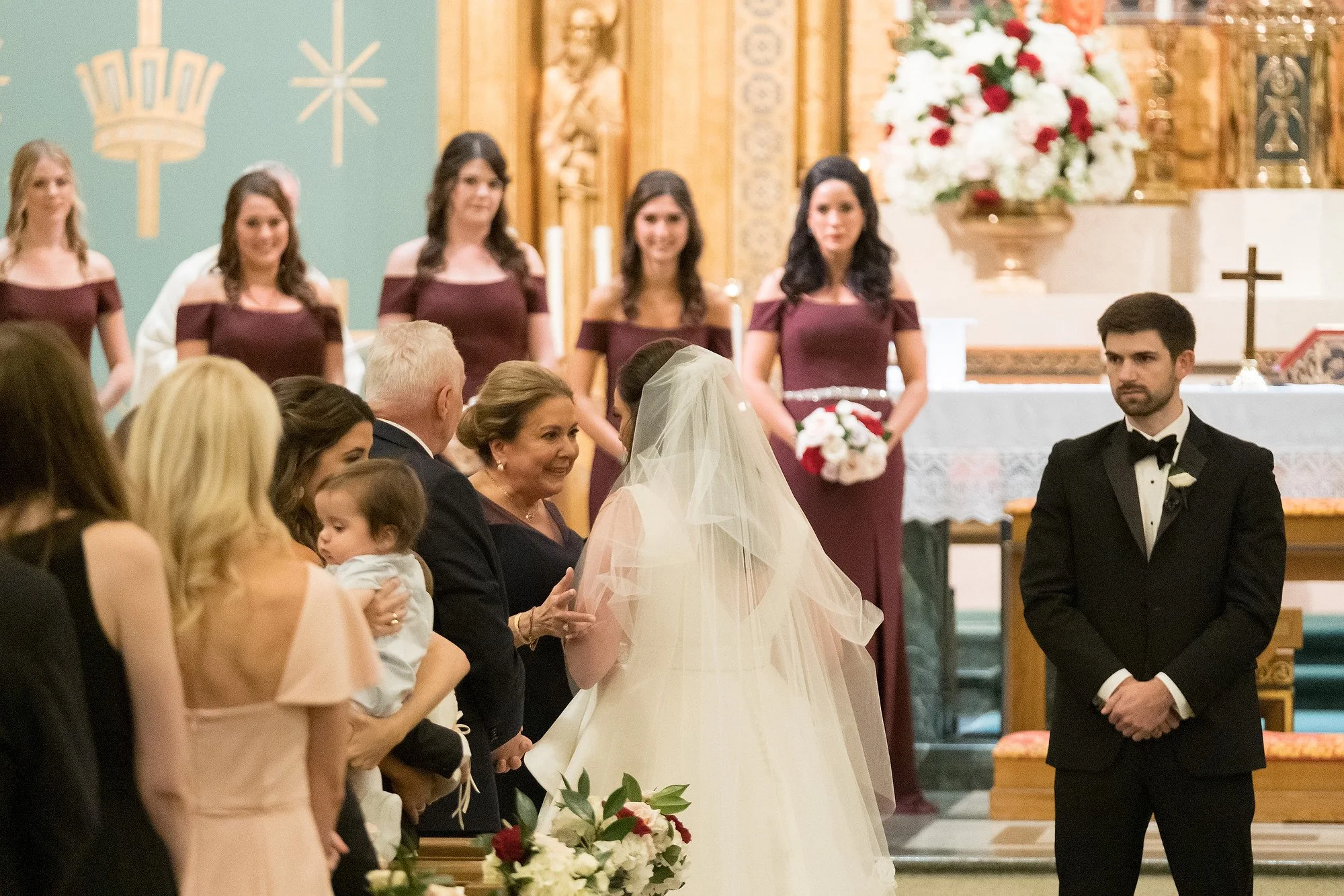 A bride in a white wedding gown and veil standing beside a woman, likely her mother, during a wedding ceremony inside a church. The groom, dressed in a black tuxedo, stands nearby. In the background, a group of women in matching burgundy dresses are 
